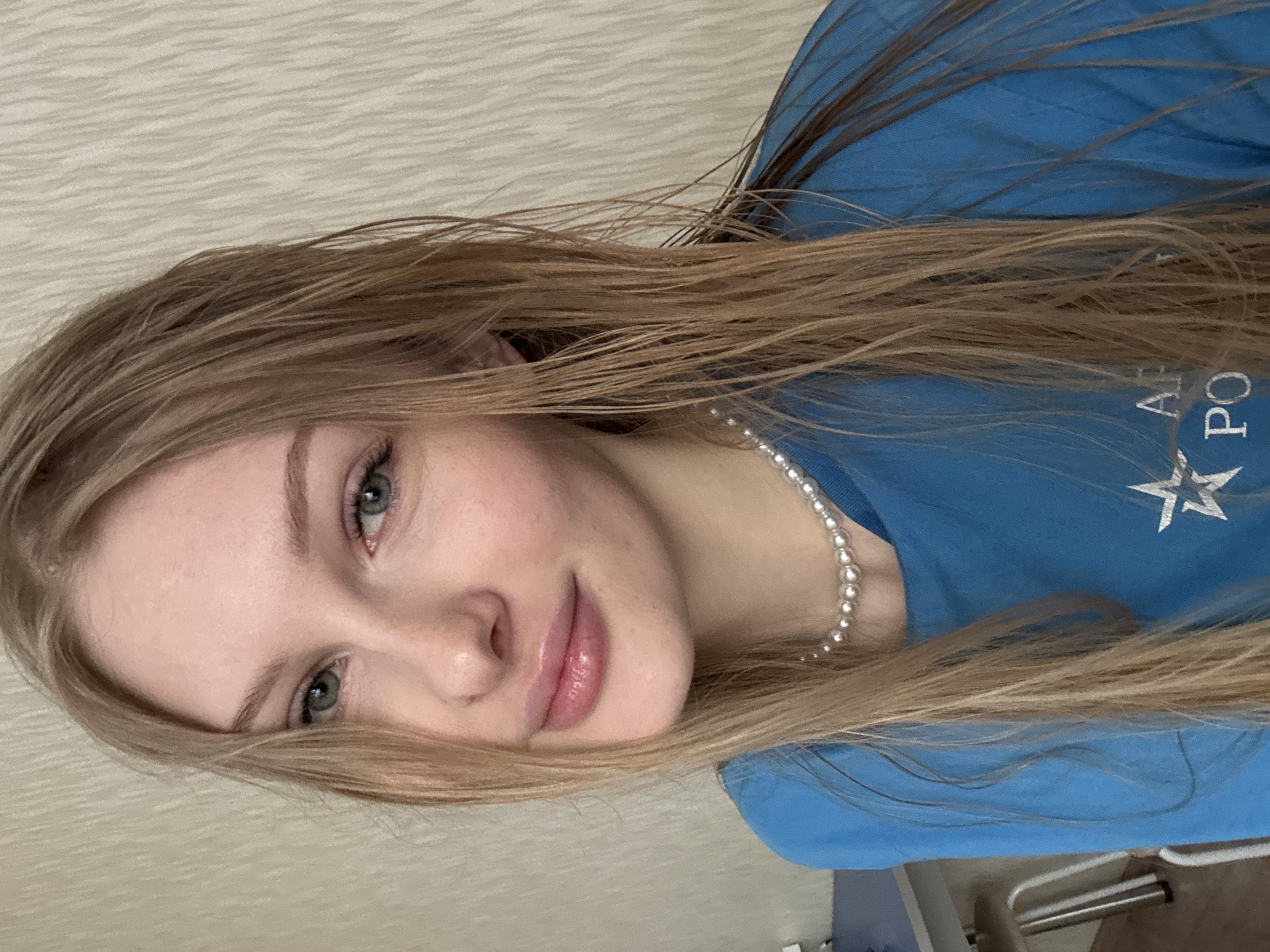 A young woman with long, straight, light brown hair and light-colored eyes (likely green or hazel) is featured in a close-up, angled portrait. Her complexion is fair, and she has a relaxed, subtle smile or pleasant expression on her face. She is wearing a light blue t-shirt and a string of pearl beads as a necklace. On the blue t-shirt, white Cyrillic text is partially visible, appearing to read "АР ПО" (likely part of "АРМИЯ РОССИИ" - "ARMY OF RUSSIA" or "АРМИЯ ПОБЕДЫ" - "ARMY OF VICTORY"), accompanied by a white five-pointed star. The background is a light-colored, subtly textured wall, suggesting an indoor setting. The image is rotated, presenting the woman's head tilted to the right. The overall impression is a casual, personal photograph taken indoors in Кондратово, Russia, under natural-looking light during the daytime.