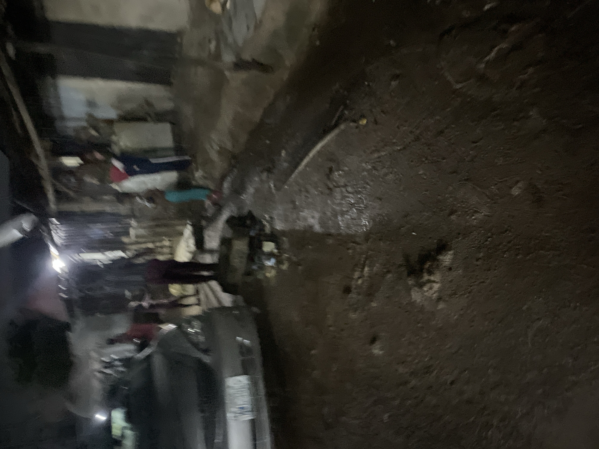 The image depicts a nighttime scene on a dirt or muddy street, possibly in a low-income area of Abuja, Nigeria. A silver sedan is parked on the left side of the frame, partially obstructing the view of a makeshift structure with corrugated metal walls. Several individuals are present. In the foreground, the ground is covered in mud and debris. In the middle ground, a group of people, including children, are visible near the structure. Some are standing, while others appear to be moving. The lighting is dim, with some artificial light sources casting shadows and illuminating parts of the scene. The overall impression is one of an unpaved, possibly underdeveloped urban environment.