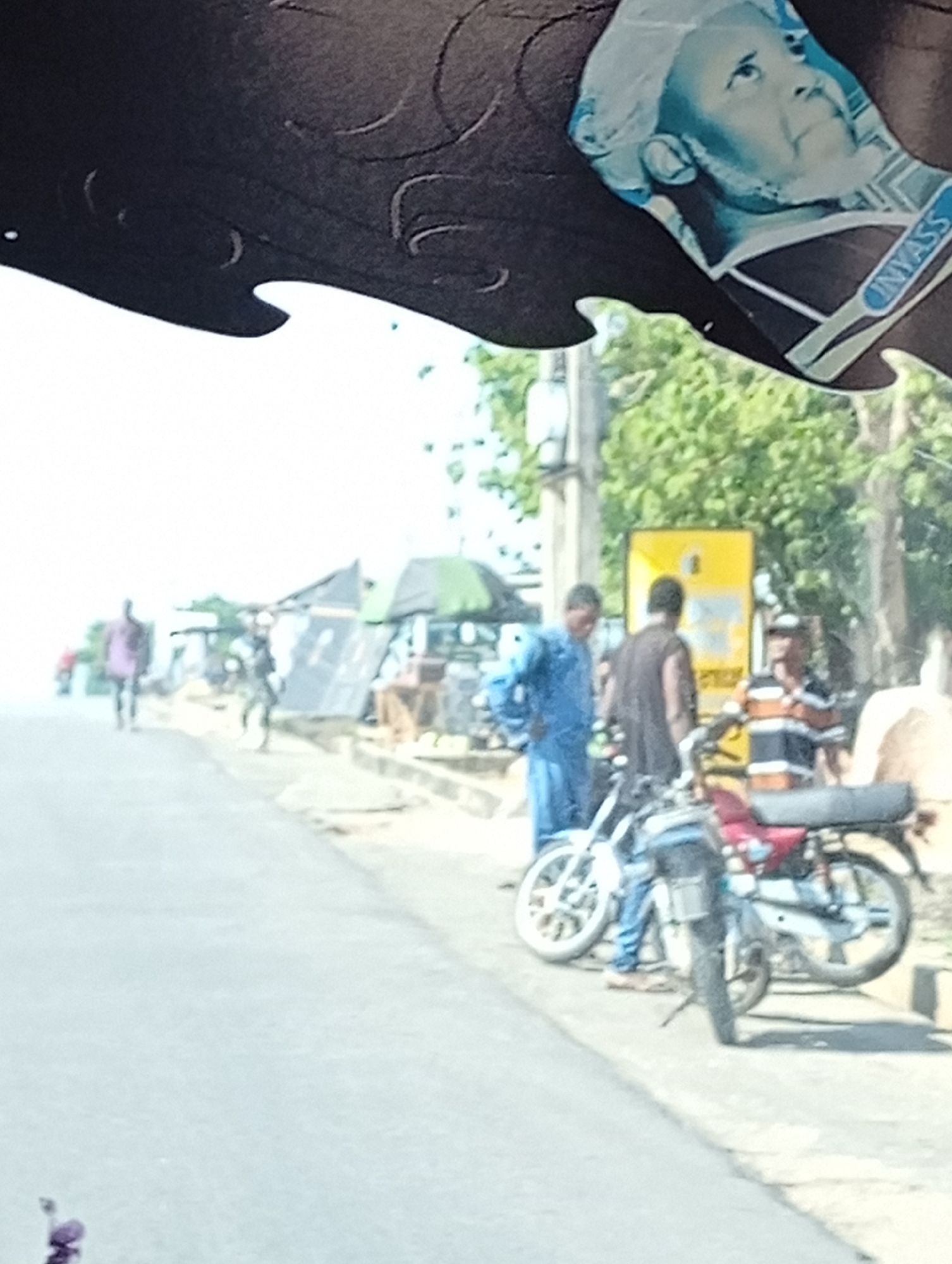 The image depicts a street scene in the city of Unknown, Unknown. Three men are standing on a sidewalk beside two parked motorcycles. One man is wearing a blue outfit, another a sleeveless black top, and the third a striped shirt. The area includes shops and stalls along the street. A figure is walking in the distance along the road. Part of a decorative dark object and a figure on currency are in the foreground, partially obstructing the view.