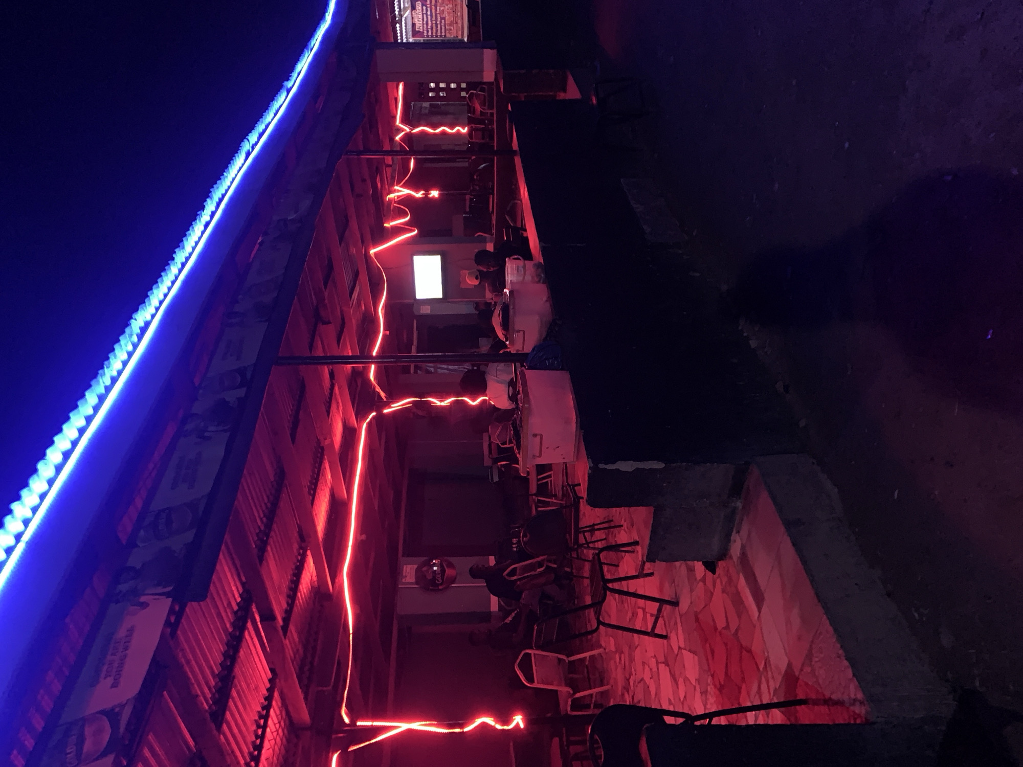 The image depicts an outdoor area in New Stadium, Nigeria, at night. A structure with a corrugated metal roof is illuminated with blue LED strip lights along its edge and red neon lights strung underneath. The area is set up as a dining or gathering space with tables, chairs, and people seated. A television screen is visible inside. The floor is tiled, reflecting the red light.
