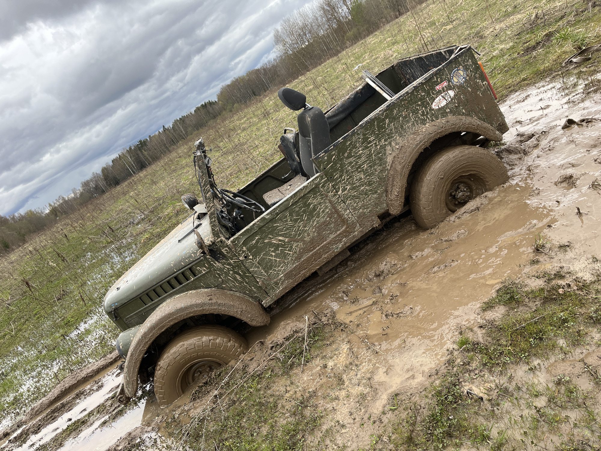 An olive green military-style vehicle is stuck in deep mud in a field in Раздолье, Russia. The vehicle has two front seats and no doors. The tires are partially submerged in thick, brown mud. Splatters of mud cover the vehicle. The field is a mix of grass and mud, with trees visible in the background. The sky is overcast with heavy, gray clouds.