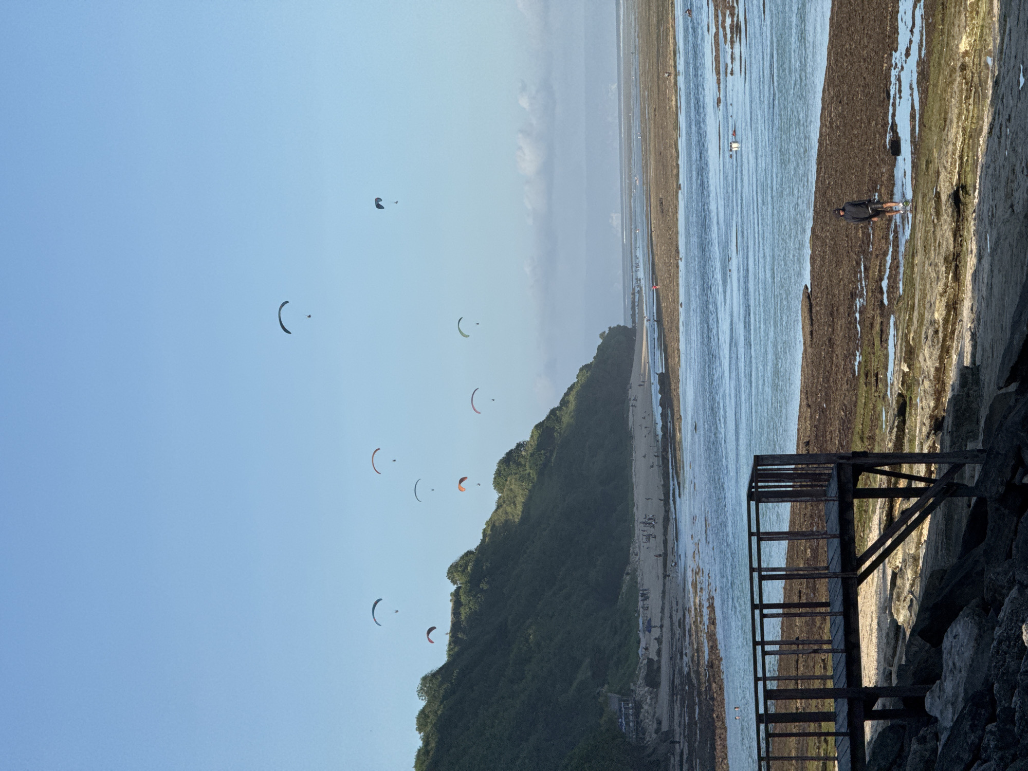 The image shows a scene in Kuta Selatan, Indonesia. Several paragliders are in the sky above a tree-covered hill that slopes down to a beach. The beach has many people on it. To the right, water meets a rocky shoreline where a person is standing. A weathered wooden structure is also visible on the rocks.
