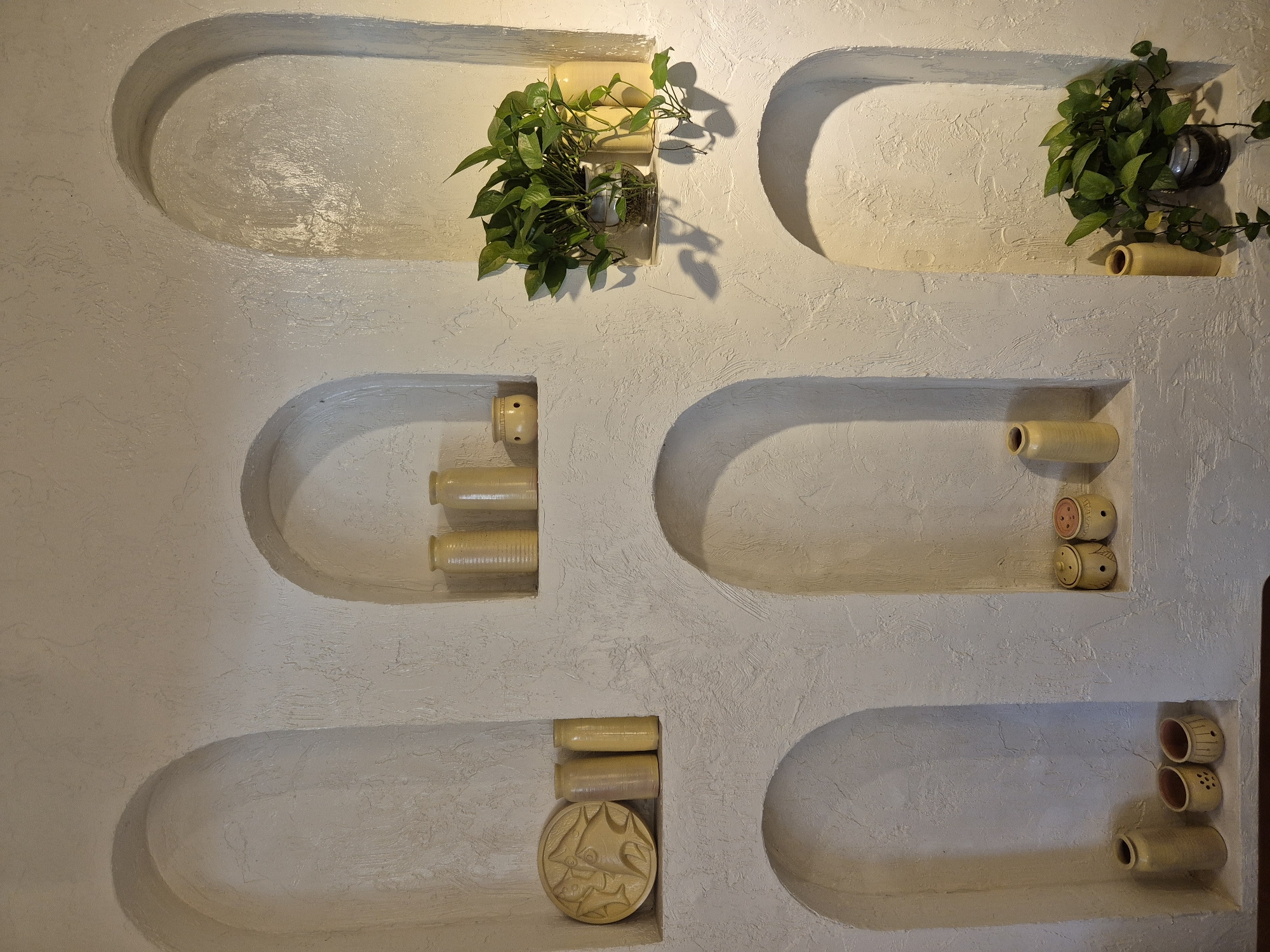 This is a close-up, eye-level shot of a textured, cream-colored wall featuring a series of recessed arched niches. The niches contain various ceramic decorative objects and a potted plant. On the top left, a decorative circular disc with raised patterns and three cylindrical vases are visible. The middle niche to the right displays three tall cylindrical vases. In the top right niche, a potted green plant in a clear glass container is present, flanked by two cylindrical vases. The bottom left niche holds one tall cylindrical vase and two small, round, lidded pots. The bottom middle niche contains two small, round, lidded pots with decorative etchings. The bottom right niche has one tall cylindrical vase and the base of a potted plant. The lighting suggests an indoor environment with warm illumination casting shadows. The scene is likely from a restaurant or home decor setting in Da Nang, Vietnam.