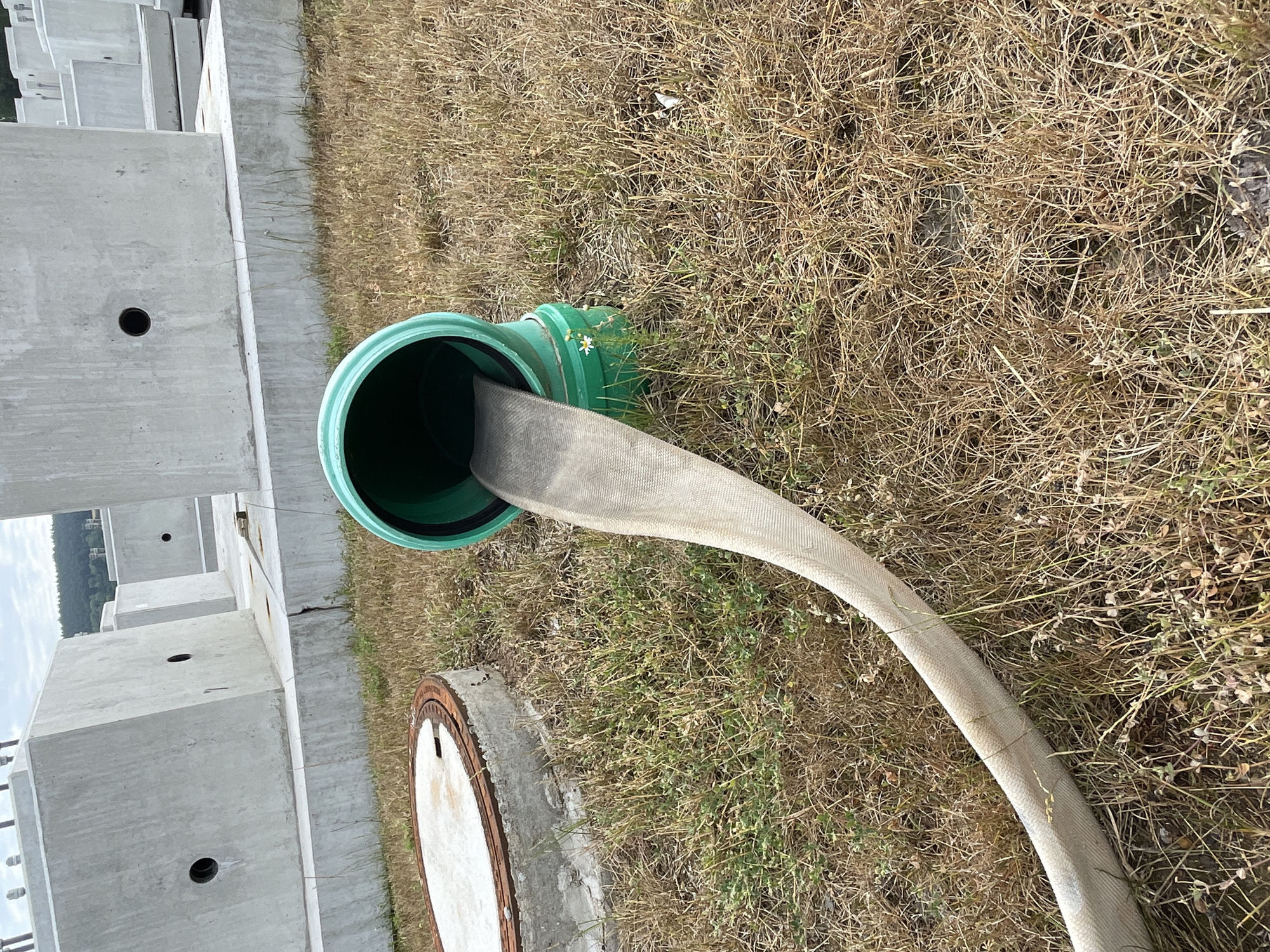 An outdoor scene in Bad Salzschlirf, Germany, depicts a construction or maintenance area. In the foreground, a flexible, light-colored hose with a woven texture extends from a green pipe fitting that is inserted into the ground. The hose lies on dry, brown grass and some sparse green vegetation. To the left of the hose, a circular concrete structure with a metal rim is partially visible, possibly a manhole cover. In the background, several large, rough-textured concrete blocks are stacked, with some showing circular openings. The sky is overcast. The scene appears to be a snapshot of ongoing work or a temporary setup at a site characterized by dry ground cover and large concrete elements.