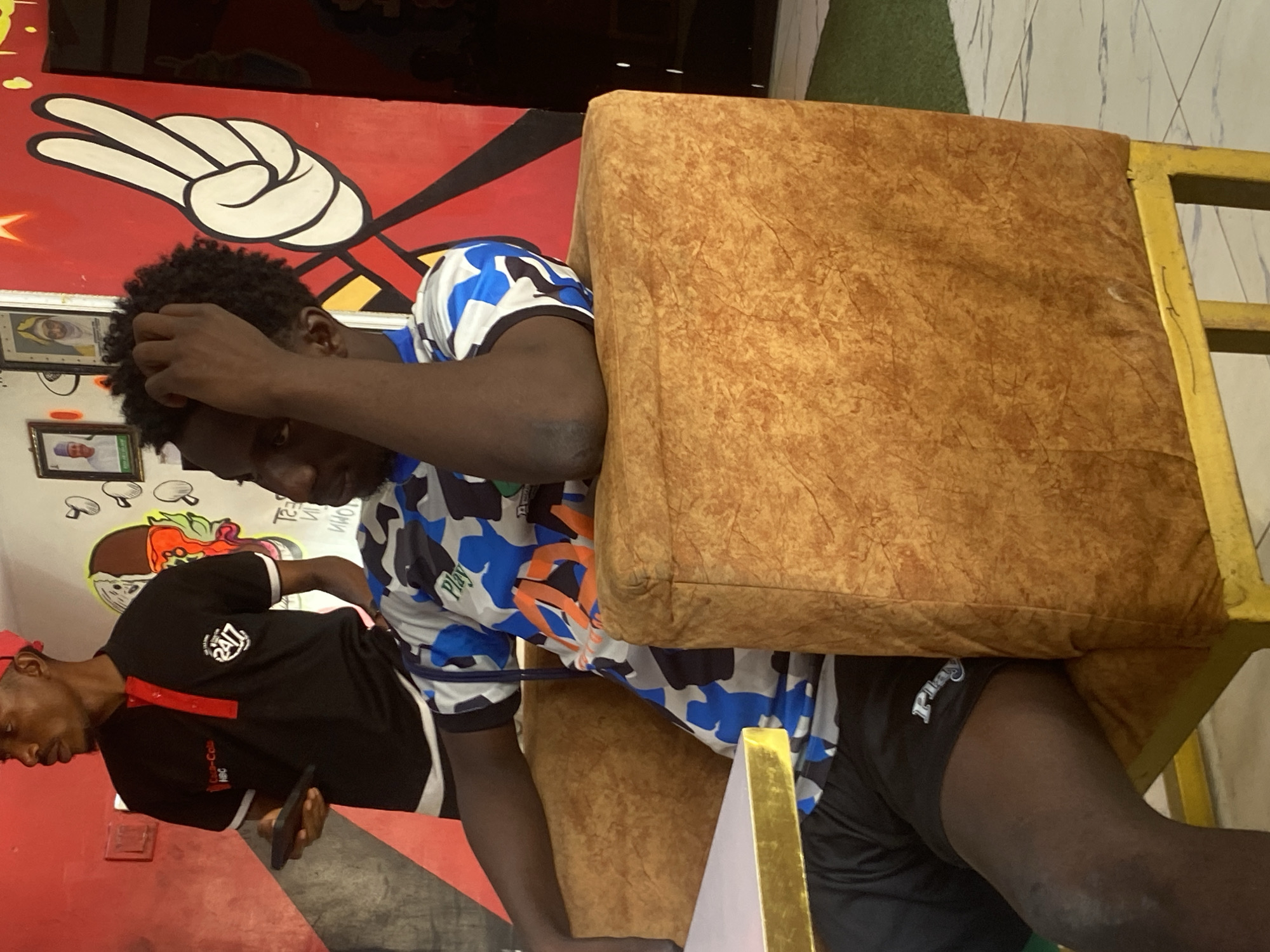 A medium shot captures two Black African men indoors against a brightly colored and patterned background. The man closer to the camera is seated, wearing a blue and white patterned shirt and black shorts with "Play" printed on them. He is leaning into a brown cushion attached to a yellow chair, his hand raised near his head as if in thought or discomfort. The second man stands to the left, wearing a black shirt with "24/7" printed on it and a red cap. He holds a phone. The backdrop includes a red wall with black lines, a white peace sign, and framed pictures. A mural featuring mushrooms and an orange-filled brown bowl, likely food-related, is also visible on the white wall. Artificial green turf is present near the top right of the frame. The image appears to be taken at eye level, with the men as the central subjects. There are no other discernible markers to conclude it is Tukur Tukur, Nigeria.