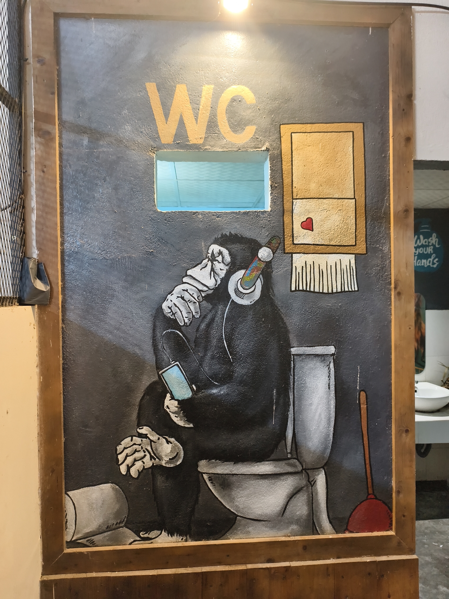The image shows a painted sign for a WC (water closet) in a restroom. A wooden frame surrounds the gray background of the sign. A cut-out rectangle provides a view of the interior. The letters "WC" are painted in gold above the cut-out. Below the letters is a painting of a chimpanzee wearing headphones and holding a smartphone while sitting on a toilet. Next to the toilet is a plunger. Above the toilet is a painting of a dispenser. The dispenser contains a red heart and toilet paper. A roll of toilet paper is on the ground near the chimpanzee. The location is Bac Nha Trang Ward, Vietnam. The backdrop beyond the restroom includes a wash basin, water dispenser, and a sign "Wash your Hands."