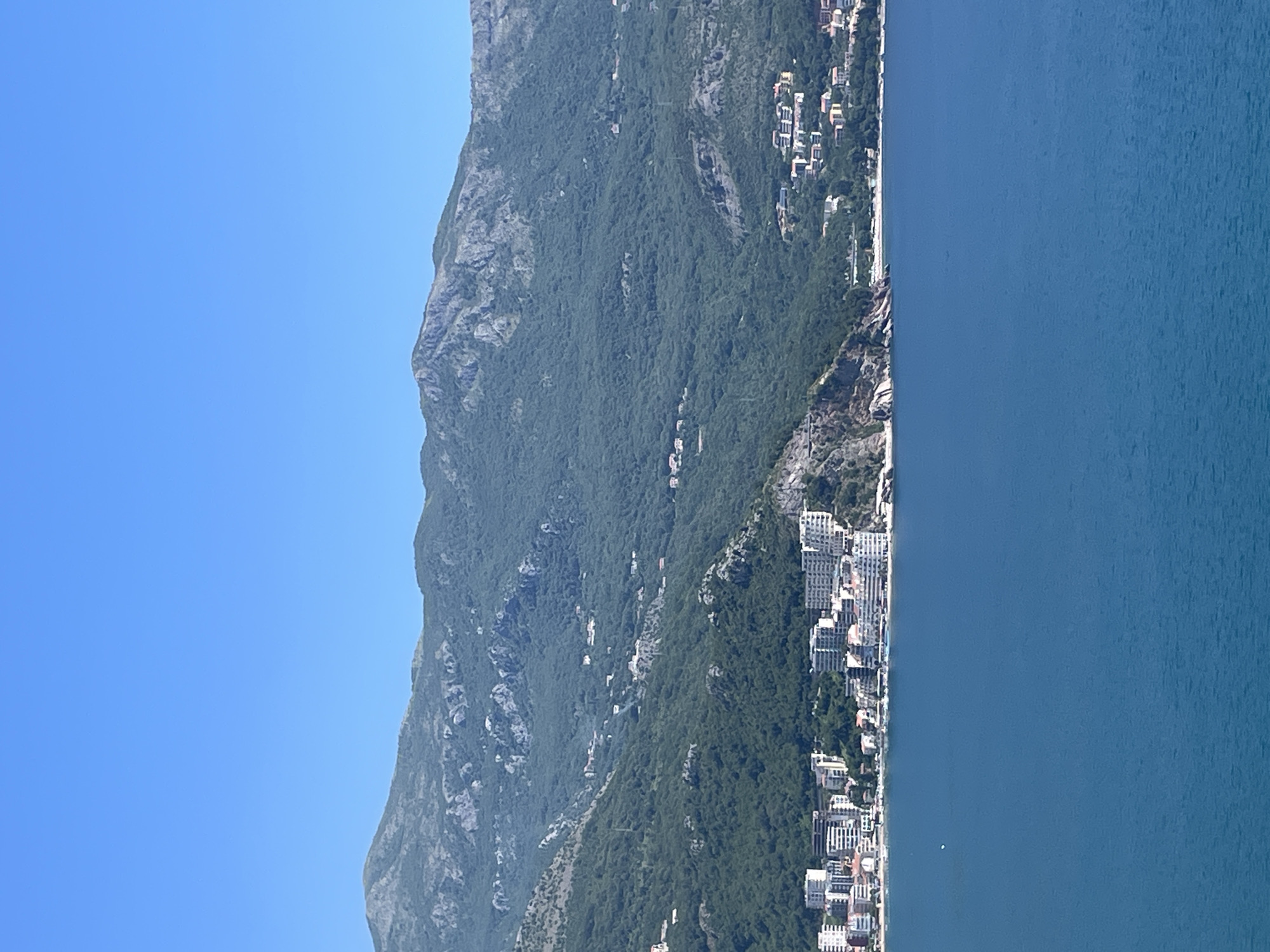 A landscape view shows a portion of the city of Boreti, Montenegro. A rugged mountain, densely covered in green vegetation, rises to the left of the frame, set against a clear blue sky. The city is situated along the base of the mountain, extending to the edge of a deep blue body of water. The city is composed of a variety of buildings, including residential homes and hotels, densely packed together, appearing white and light in color, and they stand in sharp contrast to the dark green mountain and blue water.