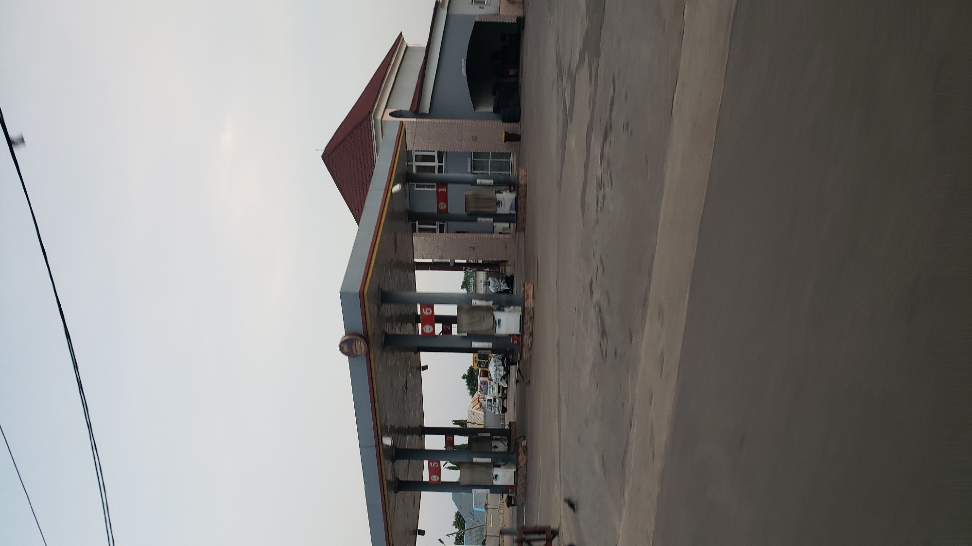 This is a daytime, eye-level, medium shot of a gas station and attached building in Jalingo, Nigeria. The gas station has a gray canopy supported by pillars. Several cars are parked at the pumps, some under the canopy. A black bird is on the asphalt in the foreground. The building behind the gas station is painted gray and has a red-tiled roof. Tires are stacked in an open bay on the right side of the building. The sky is clear and overcast.