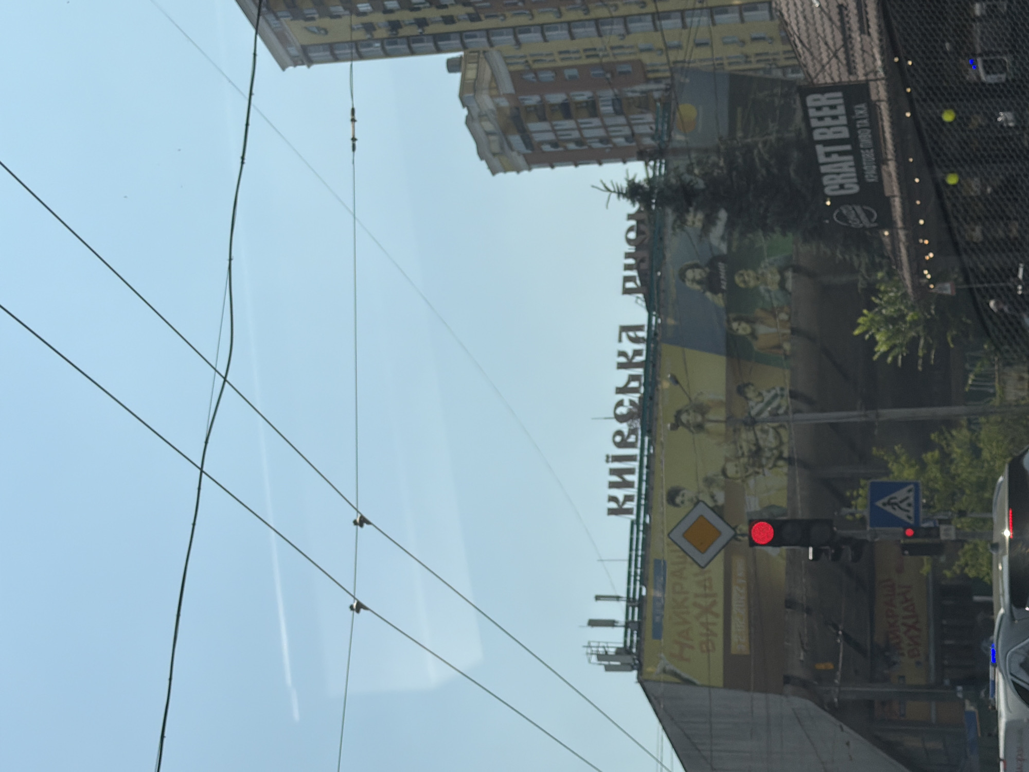 The image shows a view of a busy street in Kyiv, Ukraine, on a clear day. In the foreground, traffic lights are visible, with the red light illuminated. A pedestrian crossing sign is also present. Beyond the traffic signals, a large billboard displays images of people and Ukrainian text. The text on the billboard appears to be advertising something, possibly related to entertainment or a local event, with phrases like "НАЙКРАЩІ ВИХІДНІ" visible. Above the billboard, the words "КИЇВСЬКА РІЧКА" are displayed in large letters, which translates to "Kyiv River".
Further in the background, modern apartment buildings dominate the skyline. One tall yellow building with multiple balconies stands out, while another red-brick building is partially visible. Overhead, power lines crisscross the sky, indicating an urban setting with electrical infrastructure. A small evergreen tree is also situated near the billboard. The overall scene suggests a typical urban environment in Kyiv, with a mix of advertising, traffic management, and residential architecture.