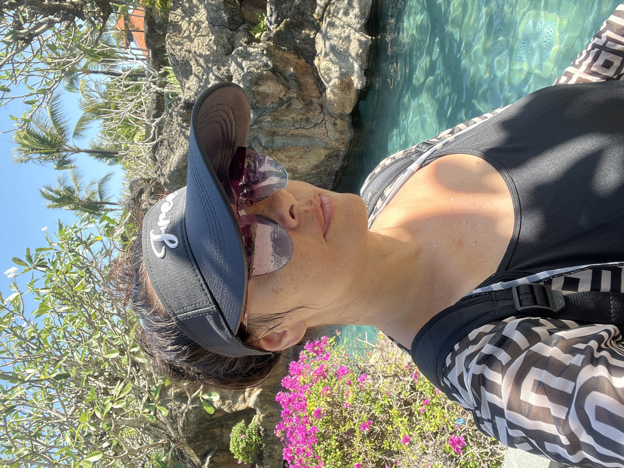 A woman stands by a swimming pool in Kuta Selatan, Indonesia. She is wearing a black visor with white lettering, sunglasses, a black swimsuit, and a black and white patterned cover-up. The pool water is clear blue, and there are rocks and lush greenery surrounding it. Palm trees and a building with an orange roof are visible in the background. The sun is shining, casting shadows and highlights on the scene.