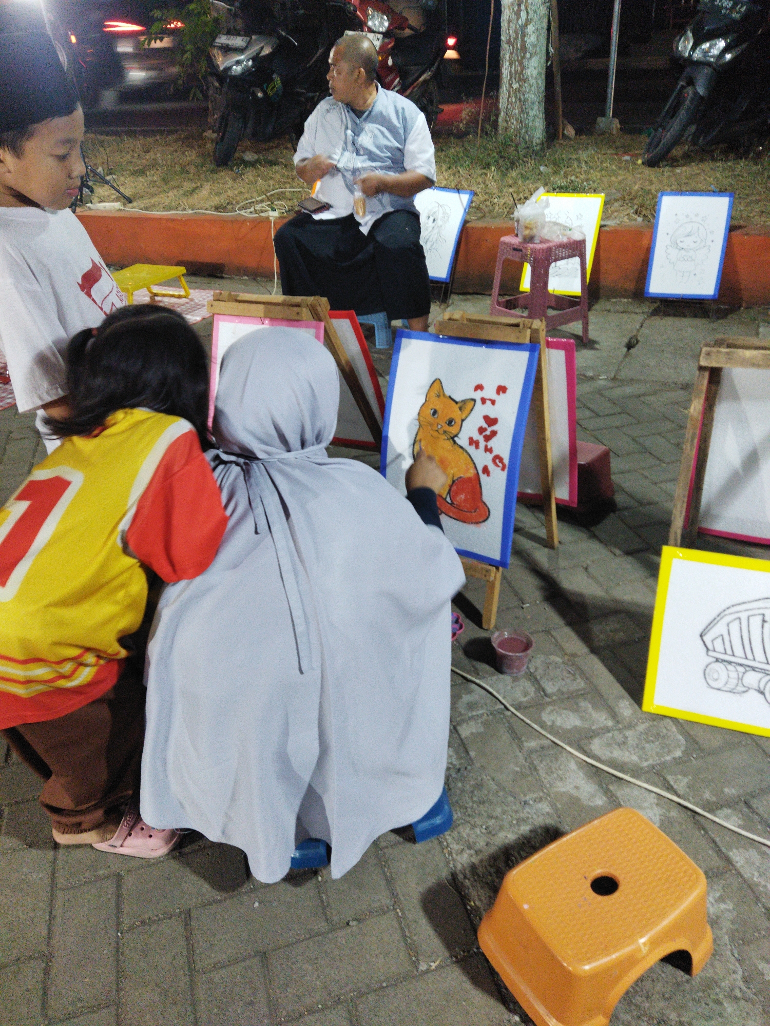 In Semarang, Indonesia, at night, a man is seated on a blue plastic stool, dressed in a light blue shirt and dark pants, with a phone in his hand. He is near a display of children's artwork, including a painting of a cat. A young boy wearing a white t-shirt with a red design is seated on the ground, looking towards the right. Beside him, a young girl in a yellow and red shirt is also on the ground, facing away from the camera. A woman in a grey headscarf is also seated on the ground, her back to the camera, appearing to be drawing or painting on an easel. The environment includes paved ground with some dry grass and trees in the background. Several easels display artwork, and there are various small stools and a bucket with what appears to be paint. Motorcycles are parked in the background, illuminated by ambient light.
