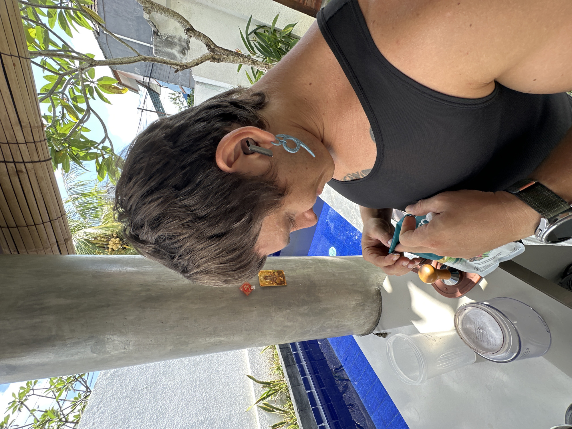 A person wearing a black tank top and a blue earring is focused on preparing something on a table outdoors. They have an earbud in their left ear and are holding a small packet and a green object. On the table, there is a clear plastic cup, a clear container with a white lid, and a coffee tamper. In the background, a large concrete pillar stands next to a blue tiled pool. The setting appears to be a tropical location, possibly North Kuta, Indonesia, with lush greenery and sunlight filtering through.