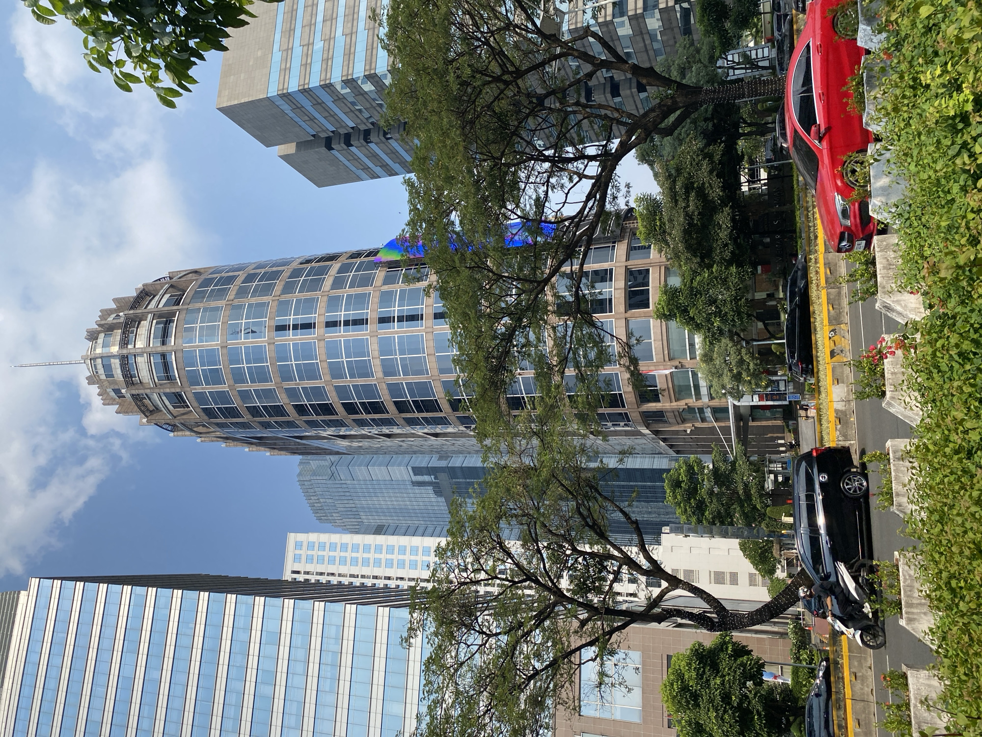 The image shows a street scene in Central Jakarta, Indonesia, with several modern skyscrapers and lush green trees. In the foreground, a red sedan is parked in the right lane of a multi-lane road, partially obscured by planters filled with dense green bushes and some flowering plants. To the left, a black minivan is driving away from the viewer, with a person on a scooter beside it. The road has yellow and white markings and a concrete divider. Behind the road, large trees with full canopies of green leaves are prominent. The background features a cluster of tall buildings with glass facades, including a distinctive cylindrical skyscraper with a decorative crown. The sky is blue with some white clouds. The overall impression is an urban environment where nature and modern architecture coexist.
