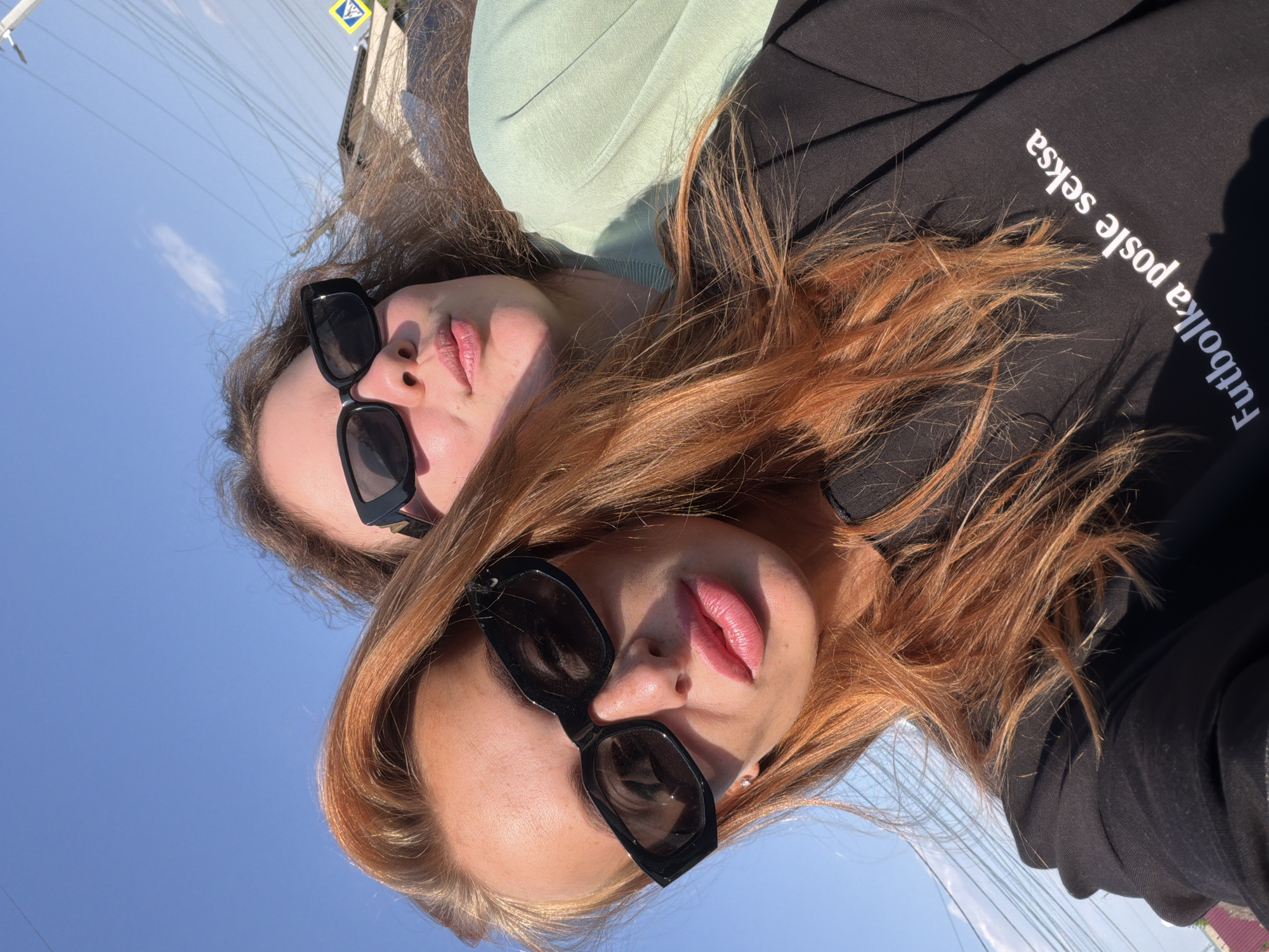 This image depicts two young adult females taking a selfie outdoors in Yemelyanovo, Russia, on a bright, sunny day with a clear blue sky. The woman in the foreground, with long reddish-orange hair, full pink lips, and a tanned complexion, wears black rectangular sunglasses and a black long-sleeved top that bears the white Latin script "Futbolka posle seksa." Her left ear features a small stud earring. Positioned behind and to her right, a second woman with wavy brown hair wears similar black rectangular sunglasses and a light green crew-neck sweatshirt, with her lips slightly pursed. The background reveals a suburban environment with numerous overhead power lines, a utility pole, the partial view of residential building roofs (one dark, one reddish), and a yellow diamond-shaped pedestrian crossing sign. Strong sunlight creates distinct shadows.