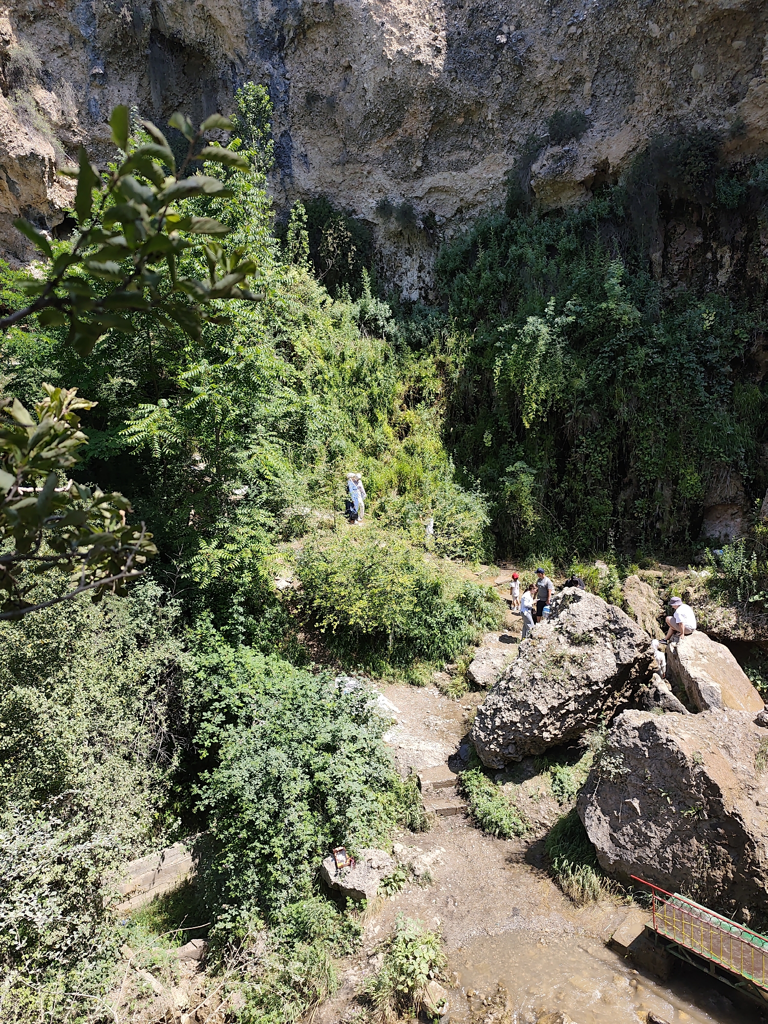 In Arstanbap, Kyrgyzstan, a natural landscape unfolds with a prominent cliff face dominating the background, dotted with vegetation. In the mid-ground, several individuals are scattered across a rocky and earthy terrain, surrounded by lush green foliage. One person in a blue shirt and white pants stands near the center, while a small group of three people, one in a red cap, are gathered on a large boulder to the left of the center. Further to the right, another individual is seated on a rock. The foreground features a muddy stream flowing beneath a small, vibrantly colored bridge with red, yellow, and green railings. The overall scene depicts a natural, outdoor setting with people enjoying the environment.