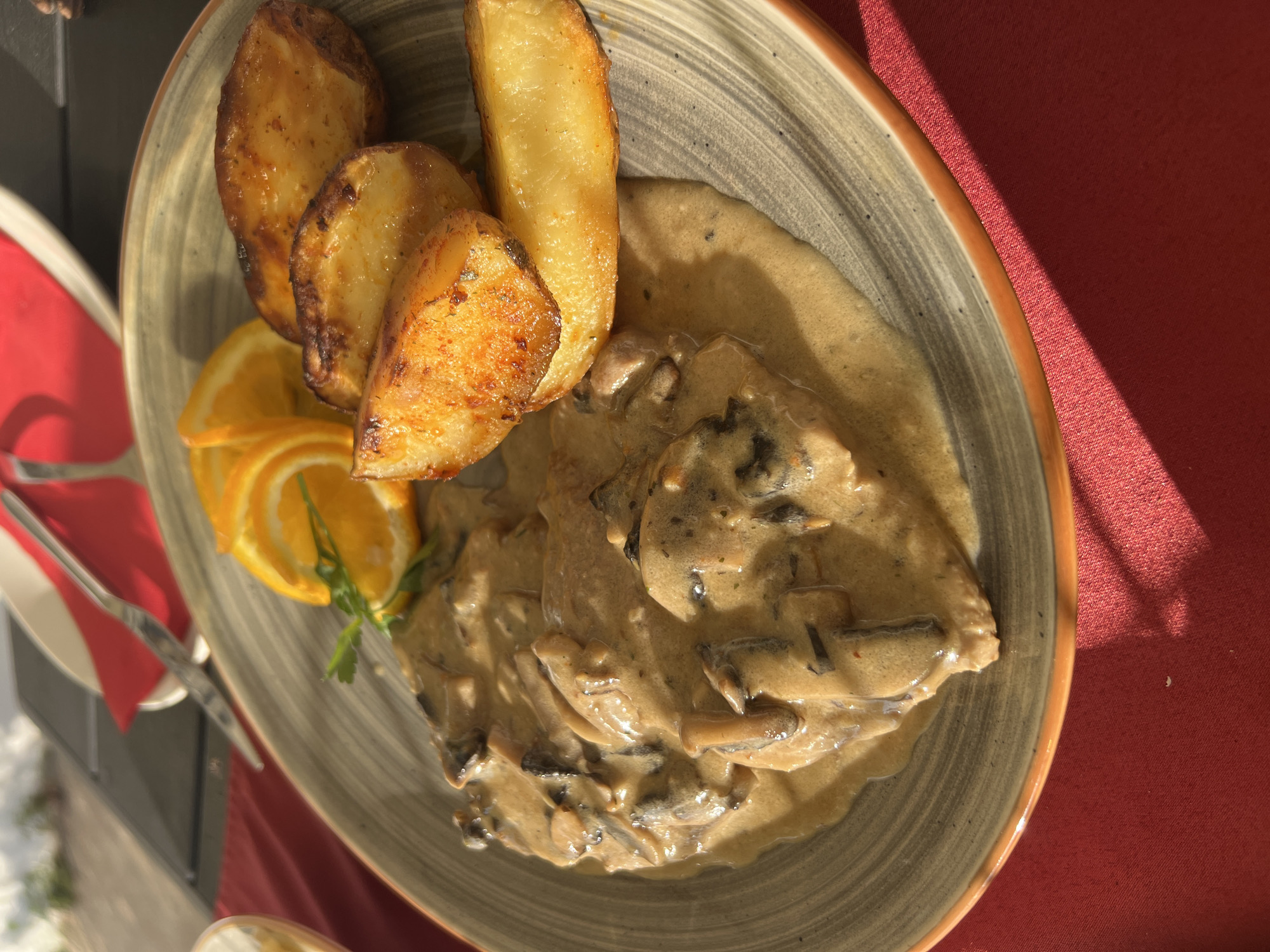 A close-up, eye-level, over-the-shoulder shot shows a plate of food on a red tablecloth. The plate, with a gray and brown striped pattern and an orange rim, contains a creamy mushroom sauce with pieces of meat, roasted potato wedges, and orange slices garnished with parsley. The scene is set outdoors, likely at a restaurant in Sveti Stefan, Montenegro, suggested by the sunlight and shadows indicating an exterior setting. The food appears to be a main course, presented in a rustic yet appealing manner.