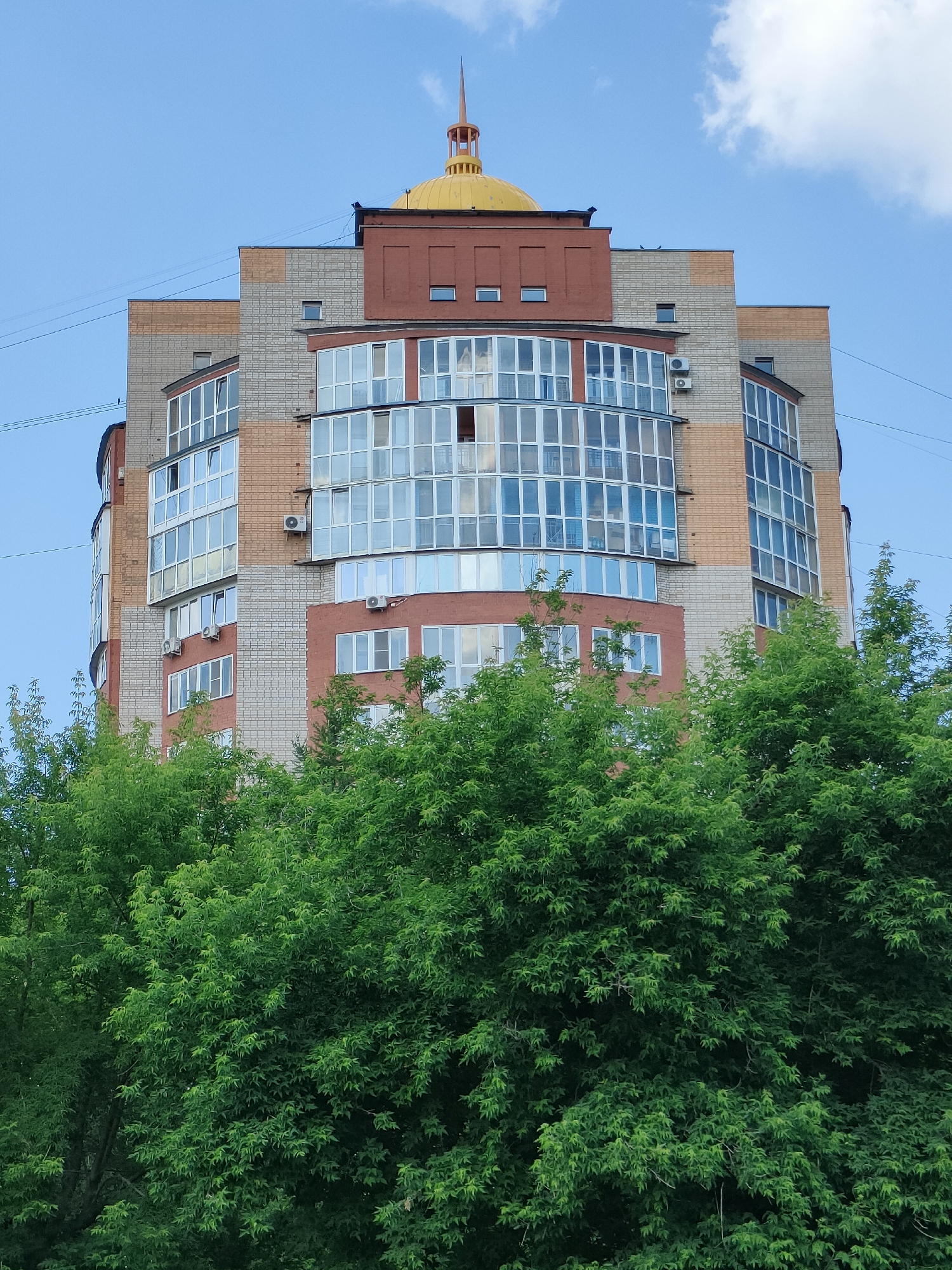 This media file is a photograph taken from a low angle looking up at a multi-story building. The building is modern in construction, with a rounded facade that features a mix of brick and what appears to be light-colored stone or concrete cladding. The windows are large and numerous, with many of them forming bay window structures that jut out from the main body of the building. Some of these windows have visible air conditioning units mounted outside them.
The building is topped with a distinctive golden dome structure, which has a spire extending from its peak. Above the dome, the sky is a clear blue with a few scattered white clouds. Power lines are visible stretching across the sky in the upper left portion of the image, suggesting the presence of urban infrastructure.
In the foreground, the lower part of the image is filled with lush green trees, their leaves creating a dense canopy that partially obscures the base of the building. The foliage is vibrant and suggests a time of year with good growth, likely spring or summer. The lighting indicates it is daytime, with the sun casting light on the building and trees. There are no people or animals visible in the image, and no text is present. The scene depicts a residential or office building set against a natural backdrop of trees, under a clear sky. The overall impression is one of a modern urban environment. The location context provided, Barnaul, Russia, situates the image within a specific geographic setting.