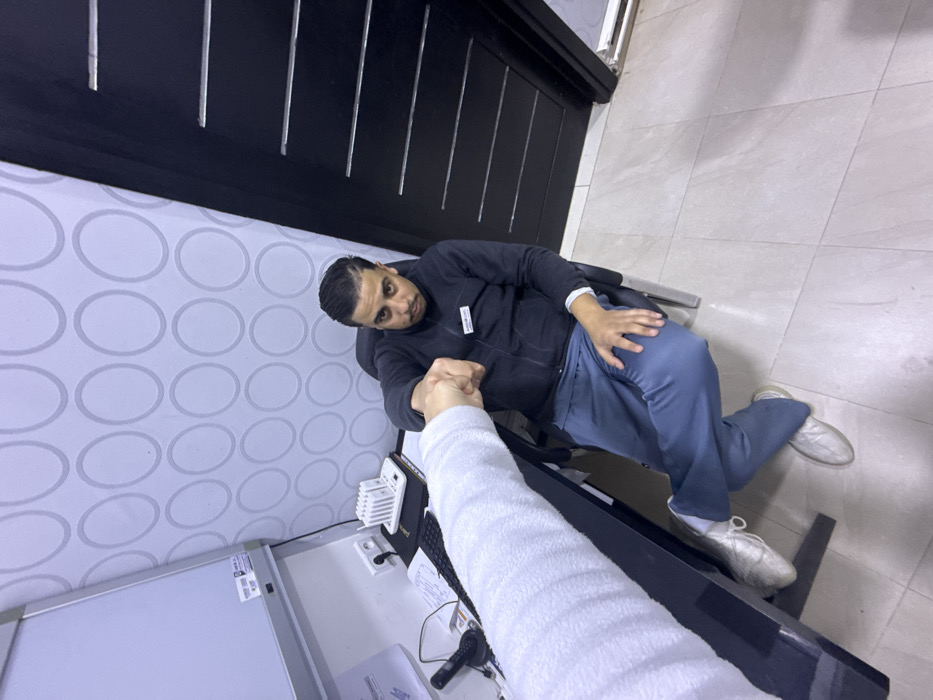 The scene depicts an indoor setting, likely an office or medical examination room, located in the city of Meknes, Morocco. The walls feature a light-colored wallpaper adorned with a repetitive pattern of light grey concentric ovals. A dark brown or black door with horizontal silver-colored accents is visible on the right, while a light-colored tiled floor covers the ground.
On the left, a dark-colored desk or counter holds various objects, including a white board or screen on a stand, a white power strip, a white electronic device with antennas, a black keyboard, and assorted papers.
A male individual is seated in a dark office chair, positioned behind the desk. He appears to be an adult with dark hair styled back and a mustache. He wears a dark blue or black zipped jacket with an ID badge on his left chest, blue or grey athletic trousers, and light-colored shoes. His left leg is crossed over his right, and his right hand rests on his left thigh.
The male individual is engaged in a fist bump with another person whose left arm, covered in a white textured sleeve, and clenched fist are visible extending from the lower left foreground into the center of the frame. The visible male's right fist meets the other person's fist, indicating an interaction. He looks directly forward with a neutral expression.