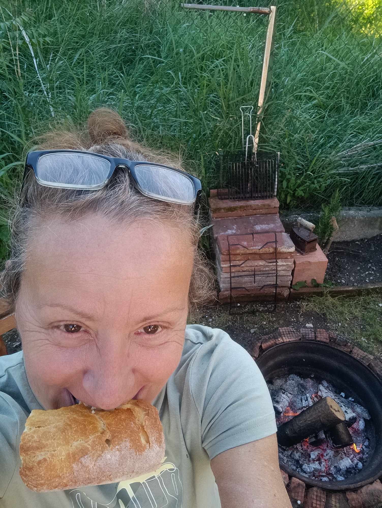 A middle-aged woman with grey hair, sunglasses on her head, and a light green t-shirt is holding a loaf of bread with melted cheese in her mouth. Behind her is a makeshift grill made of bricks with a grilling rack on top, and to the right is a fire pit with burning wood and embers. The location appears to be outdoors, with tall green grass in the background. This is possibly located in the city of Viadana, Italy.
