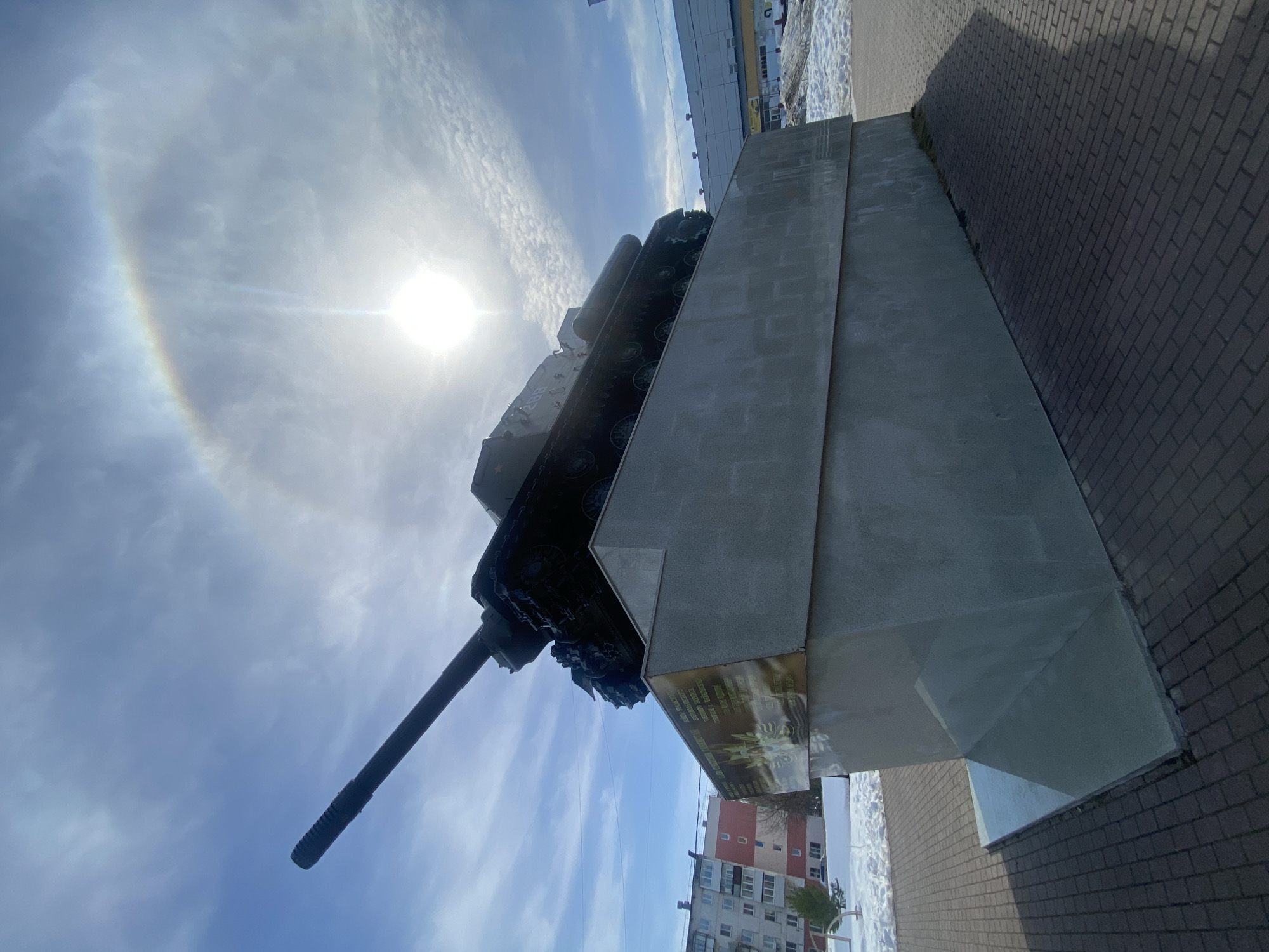The image shows a tank mounted on a concrete plinth, likely a war memorial. The tank is positioned with its gun pointing diagonally upward towards a bright sun, surrounded by a circular rainbow (halo). The plinth is multi-tiered and appears to have a plaque affixed to one side. The background includes a brick-paved area, a building with windows, possibly residential, and another larger commercial-looking structure. The photo is taken at an angle, giving the impression the tank is tilted. Based on information from context, the scene is located in the city of Chaikovsky, Russia. There are no people visible in the image.