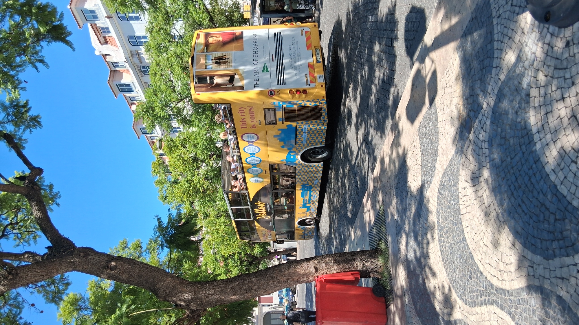 This is a full-shot, outdoor, eye-level, vertical photograph of a yellow double-decker tour bus on a sunny day in Lisbon, Portugal. The bus, branded "Yellow Bus Tours", is parked on a cobblestone street lined with trees and historic buildings. People are seated on the upper deck of the bus, enjoying the sights. The bus has an advertisement on the back that reads "THE ART OF SHOPPING". A red trash bin is located on the left side of the frame, near a tree. The sidewalk features a distinctive wave pattern made of dark and light cobblestones.