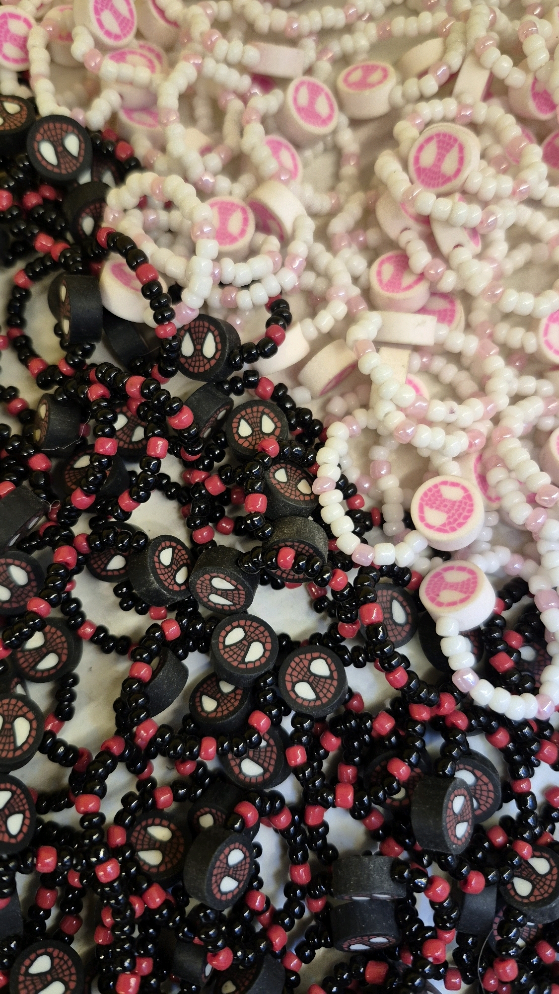 In Watuagung, Indonesia, a close-up overhead shot reveals a large pile of beaded bracelets. Two distinct styles are visible: one set features black and red beads with circular charms depicting the Spider-Man logo, and the other is composed of white and pale pink beads with similar Spider-Man logo charms. The bracelets are jumbled together, creating a textured and colorful display. The background is out of focus, but appears to be a light-colored surface. There are no people or animals present in the image.