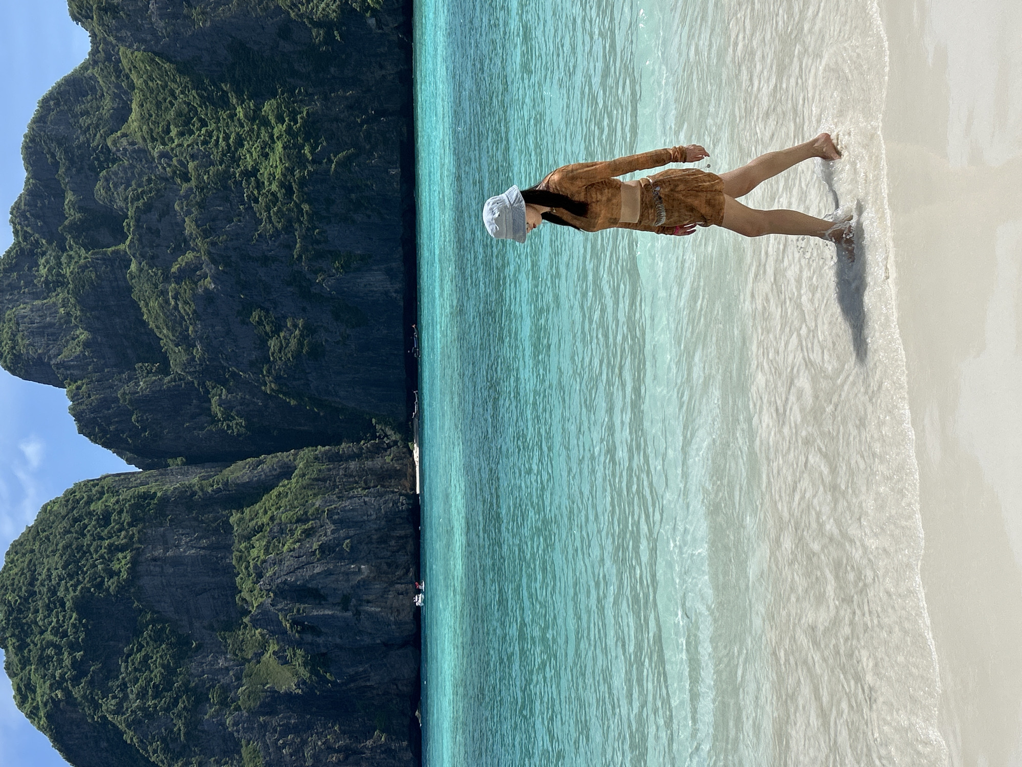 A woman wearing a light blue bucket hat and a brown two-piece outfit, with a long-sleeved top and a skirt, walks along the shoreline of a tropical beach. The clear, turquoise water laps gently at her feet as she walks towards the right side of the frame. In the background, towering, lush green limestone karsts rise dramatically from the sea, partially obscured by haze. Several small boats are visible in the distance, dotting the calm waters. The beach is composed of fine, white sand, with the waves creating a foamy white edge as they recede. The sky above is a bright blue with a few scattered white clouds. This scene is characteristic of Maya Bay, located near Ao Nang, Thailand.