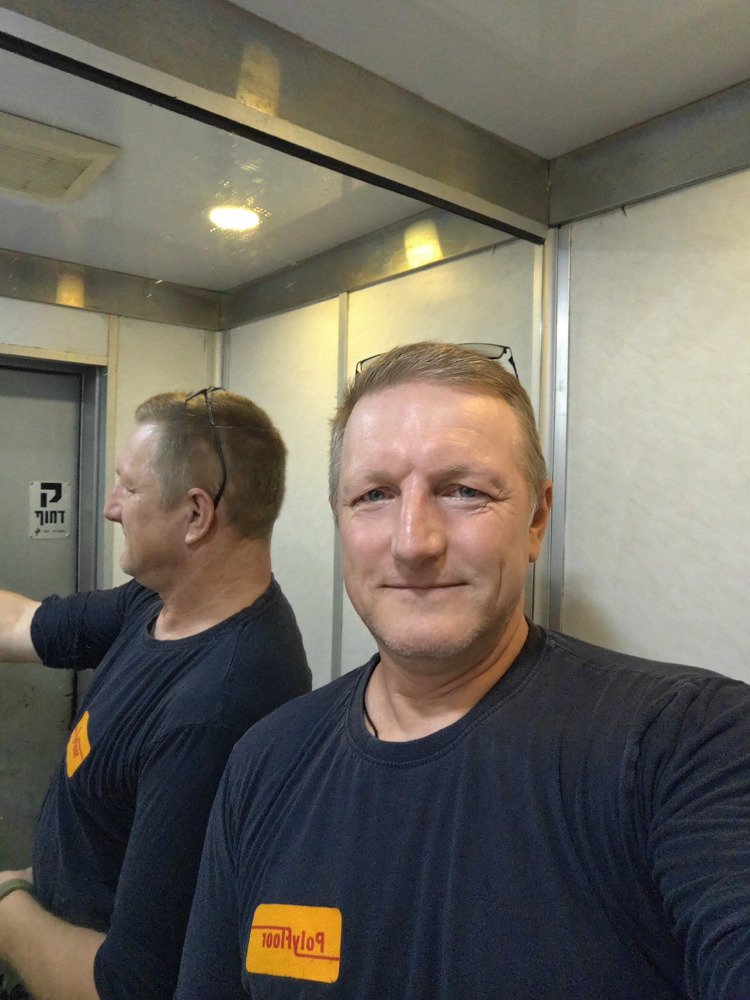 A medium shot photo shows a middle-aged Caucasian man taking a selfie in an elevator. The man has short, light brown hair, blue eyes, and a light complexion. He is wearing a dark blue t-shirt with a yellow rectangular logo on the left chest that reads "PolyRoo". He has a pair of eyeglasses perched on top of his head. The elevator interior is metallic with some white panelling and a reflective ceiling. A mirror on the left wall reflects another view of the man, showing his profile. The elevator doors are visible on the left. There is a sign on the elevator doors with Hebrew writing. The overall impression is that this image was taken in a public building, possibly an office or commercial space in the city of Petah Tikva, Israel.