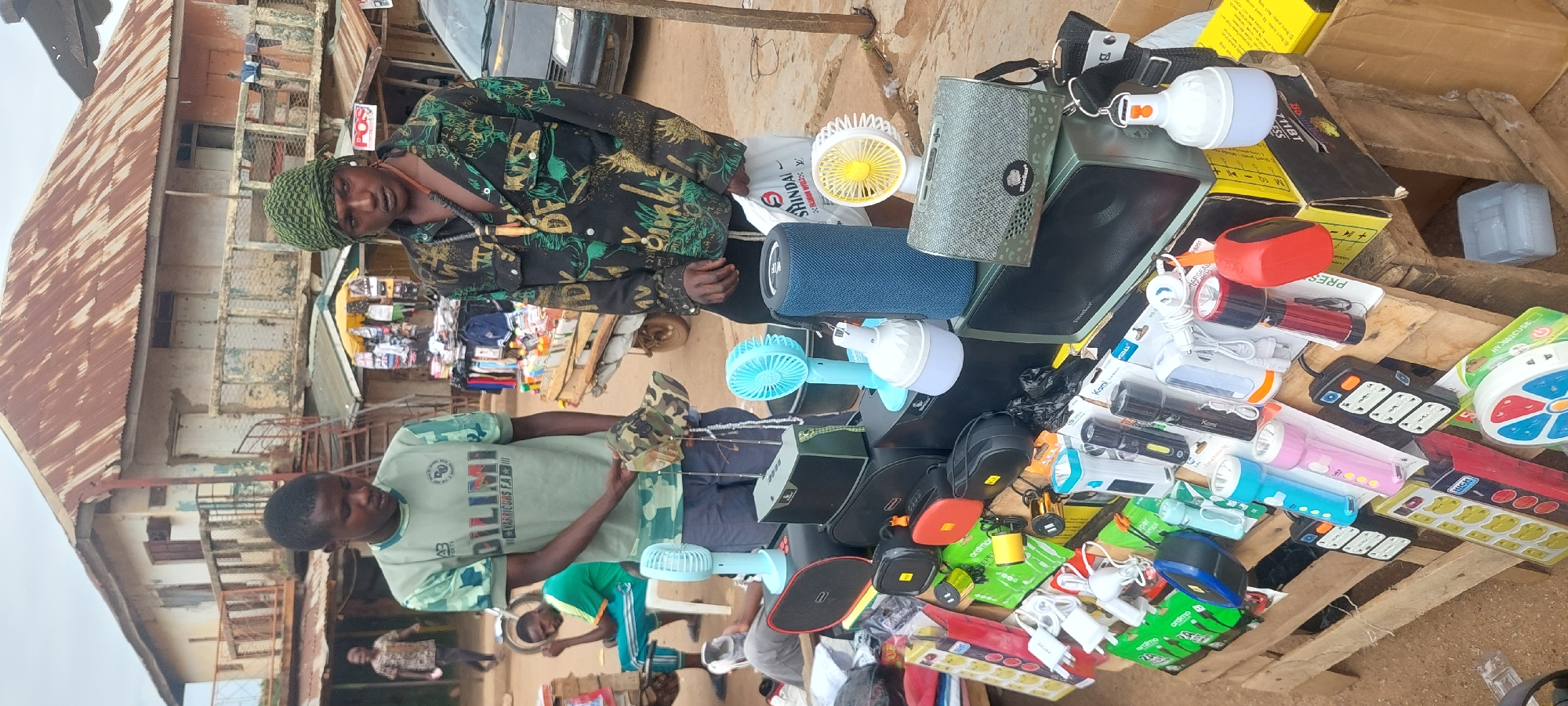 An outdoor market stall in the city of Unknown, Unknown, features two men presenting electronic goods and accessories. The scene is set on uneven, light brown dirt or gravel ground, with weathered buildings in the background.
The man on the left, a young adult with dark skin and short dark hair, wears a light green short-sleeved t-shirt with a camouflage pattern on the shoulders and sleeves, bearing text "DILIMI" and "VARRIO.F.A". He is holding up a camouflage baseball cap with both hands, inspecting it or displaying it to an unseen party, with his gaze directed downwards.
The man on the right, also a young adult with dark skin, wears a green patterned headscarf and a long-sleeved dark shirt with abstract green and gold patterns. A dark beaded necklace with a wooden pendant hangs around his neck. He stands with his right hand holding a white plastic bag with red "SHINDAI" lettering. His attention appears directed towards the left.
The market stall consists of various electronic items and small gadgets displayed on two stacked wooden pallets. These items include:
* Numerous packaged USB wall chargers and cables.
* A variety of packaged LED flashlights in colors such as blue, pink, black, and red.
* Several multi-socket power strips and extension cords, some in vibrant colors and patterns.
* Multiple small, portable battery-operated fans in light blue and yellow.
* Various portable Bluetooth speakers, including a blue cylindrical one and a grey rectangular one, along with larger black speaker units.
* White, bulb-shaped portable LED lamps with hooks.
* Black and colored pouches, possibly for electronics.
A light blue plastic container is visible beneath the display.
In the background, a large, two-story building with a rusted corrugated metal roof dominates the view. The building exhibits peeling, faded light-colored paint (cream and light orange sections) and a dilapidated balcony with a metal railing. A spiral metal staircase is affixed to the left side of this building. Smaller market stalls with hanging merchandise, including one with a "POS" sign, are also visible. A dark-colored vehicle is partially seen on the far right. Further back on the left, another man in a green t-shirt and light blue pants sits on a white plastic chair, while another individual's legs are visible lying on the ground near the stall.