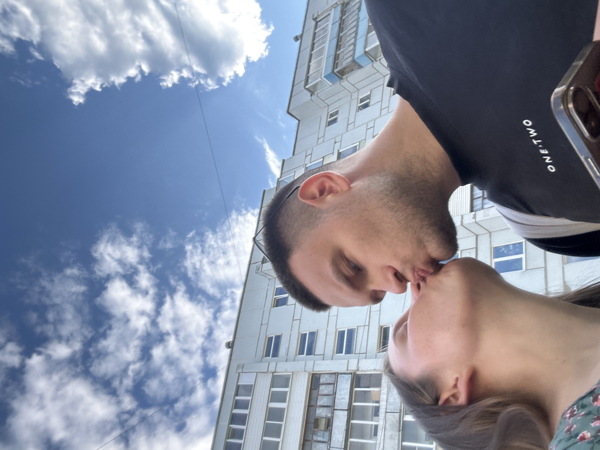 Two young adults, a male and a female, are depicted in a low-angle, close-up shot, engaged in a kiss. The female, positioned on the left, has long brown hair and is wearing a green top with a floral pattern. The male, on the right, has short dark hair, light facial stubble, and wears a black t-shirt with the white text "ONE TWO" visible on the chest. He is holding a dark-colored smartphone, possibly an iPhone, in his right hand at the bottom right of the frame.
The background features a large, light-colored, multi-story residential building, characteristic of post-Soviet era architecture, with numerous windows and balconies. Some windows on the left side display older enclosures and an external unit resembling an air conditioner. The upper portion of the image reveals a clear blue sky with scattered white cumulus clouds, and thin overhead wires traverse the sky horizontally. The scene is located in the city of Chaikovsky, Russia.