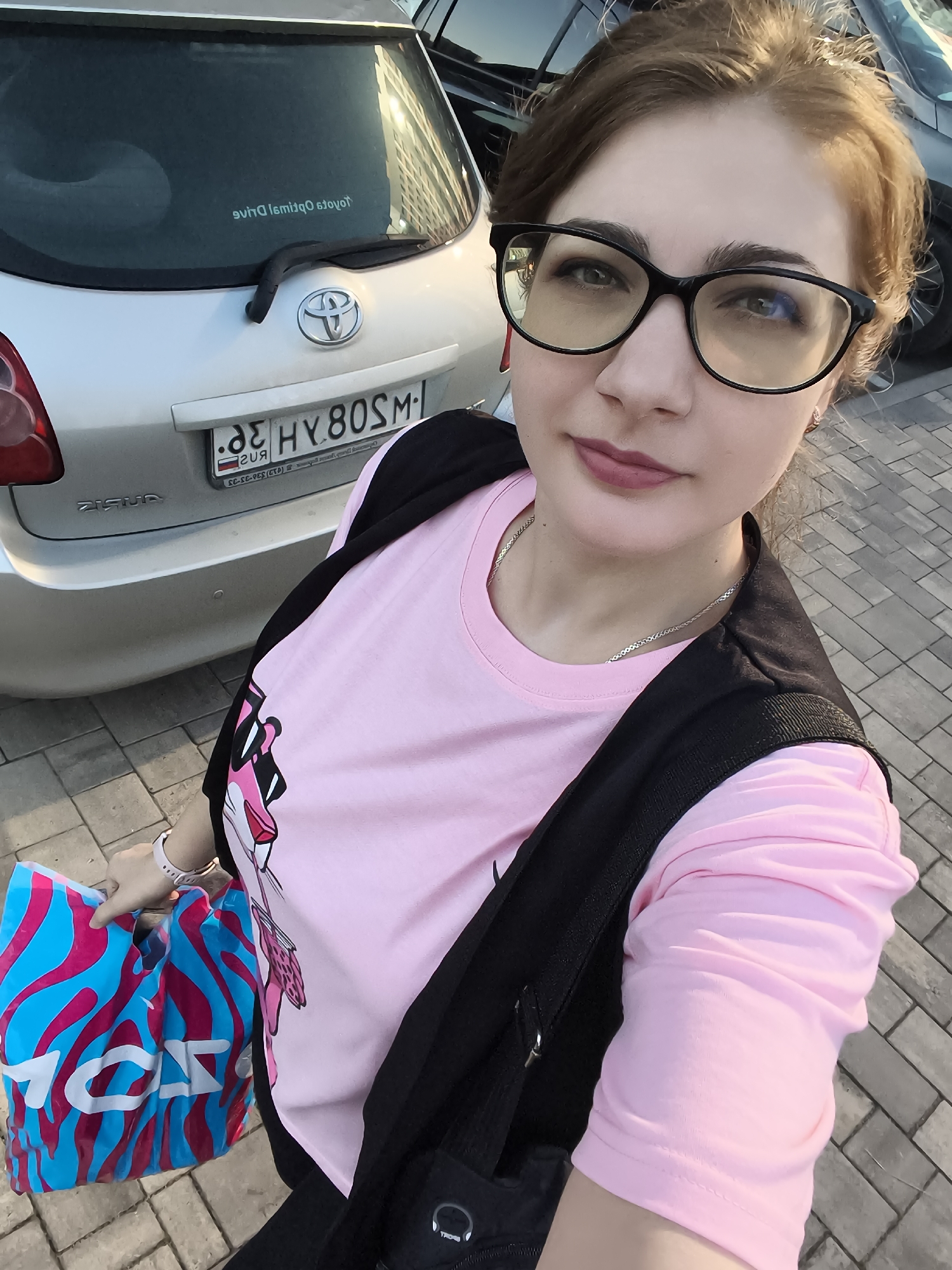 A young woman with light brown hair and glasses takes a selfie outdoors in front of a silver Toyota car. She is wearing a pink t-shirt with a graphic of a pink panther on it and a black vest or jacket draped over her shoulders. She is holding a blue and pink striped shopping bag with white lettering in her left hand. The car's license plate is visible and reads "32 HY 80SM". The background shows a paved walkway and other cars. The lighting suggests it is daytime.