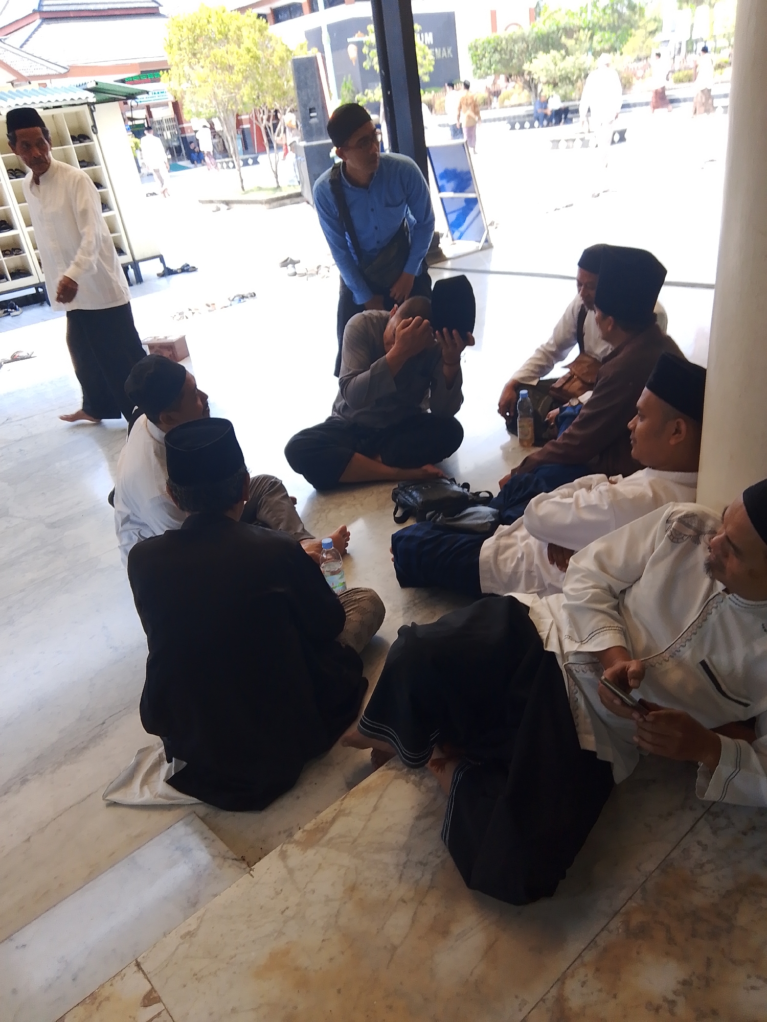 The image depicts a group of men wearing traditional Islamic attire, including peci caps and long robes, gathered in what appears to be the courtyard or entrance area of the Great Mosque of Demak, Indonesia. The architecture visible in the background, with its pitched roofs and wooden elements, is characteristic of traditional Javanese mosques. Several men are seated on the marble floor, engaged in conversation or resting. One man in the foreground is looking at his phone, while others are observing their surroundings or each other. The presence of a blue water bottle suggests a pause or break. The overall atmosphere is relaxed and informal, with the men appearing to be members of a congregation or visitors to the mosque. The lighting indicates it is daytime, with bright sunlight casting shadows.