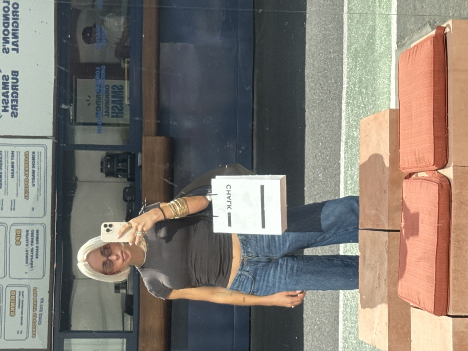 A platinum blonde woman is captured in a full-body selfie reflection, standing outdoors in front of a food establishment identified as "London's Original Smash Burgers" in Dubai, United Arab Emirates. The woman wears a dark grey crop top exposing her midriff, loose-fitting light-wash blue jeans, and amber-tinted rectangular sunglasses. Her right hand holds a light-colored smartphone, positioned to take the photo, displaying rings on her fingers and multiple gold-toned bangles on her wrist. A black shoulder bag hangs on her left side, and she carries a white paper shopping bag with black handles, featuring a black horizontal stripe and the word "CHALK" in reversed text, indicative of a reflection.
Behind the woman, the reflection shows a dark blue building facade with a large window. Above the window, white menu boards with black text display items such as "DRINKS," "DIPS," "HOT SMASH," "CLASSIC SMASH," "ALEXIA SPECIAL," and "KIMCHI MAYO." The establishment's name, "LONDON'S ORIGINAL SMASH BURGERS," is visible, partially reversed, on the window. Through the window, inside the building, a dark-colored coffee machine sits on a counter, and a person wearing a dark baseball cap and a light-colored shirt is visible further back.
The ground beneath the woman's reflection features a dark grey paved section leading to a lighter green paved area. In the immediate foreground, a structure of stacked reddish-brown brick blocks serves as seating, topped with two square orange fabric cushions. The scene is brightly lit, with shadows cast by the woman and the seating structure.