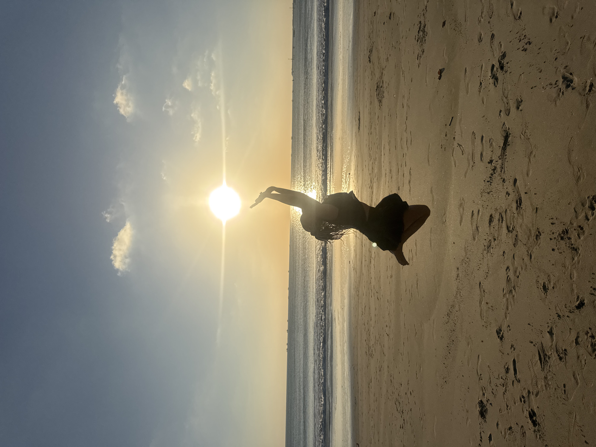 A silhouette of a person is captured mid-air, jumping on a beach at sunset. The person is wearing dark clothing and their arms are raised, with one hand reaching towards the sun, which is positioned in the upper center of the frame, low on the horizon. The sun casts a bright, warm glow across the sky, which transitions from a light blue at the top to a soft orange near the horizon. There are a few scattered clouds in the sky. The ocean stretches out to the horizon, with gentle waves breaking on the shore. The beach in the foreground is sandy and shows the imprints of footprints. The scene appears to be in Kuta, Indonesia, a popular tourist destination known for its beaches and sunsets.