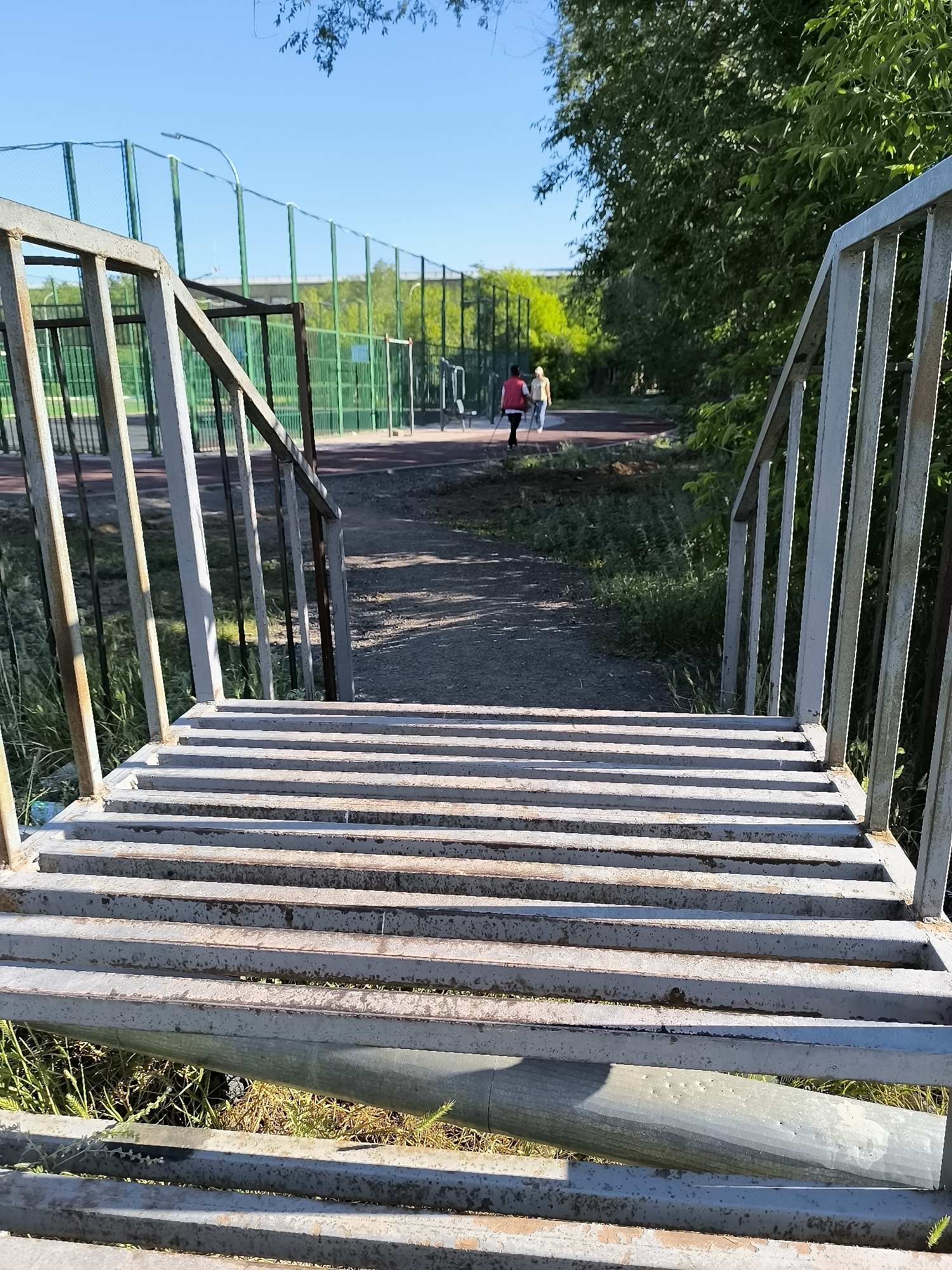 An outdoor scene in Qarağandı city administration, Kazakhstan, features a metal staircase leading to a pathway. Two individuals, one wearing a red jacket and the other a yellow top, are seen walking on the pathway. The pathway is bordered by a fenced sports field and lush green trees. The sky is clear and blue.