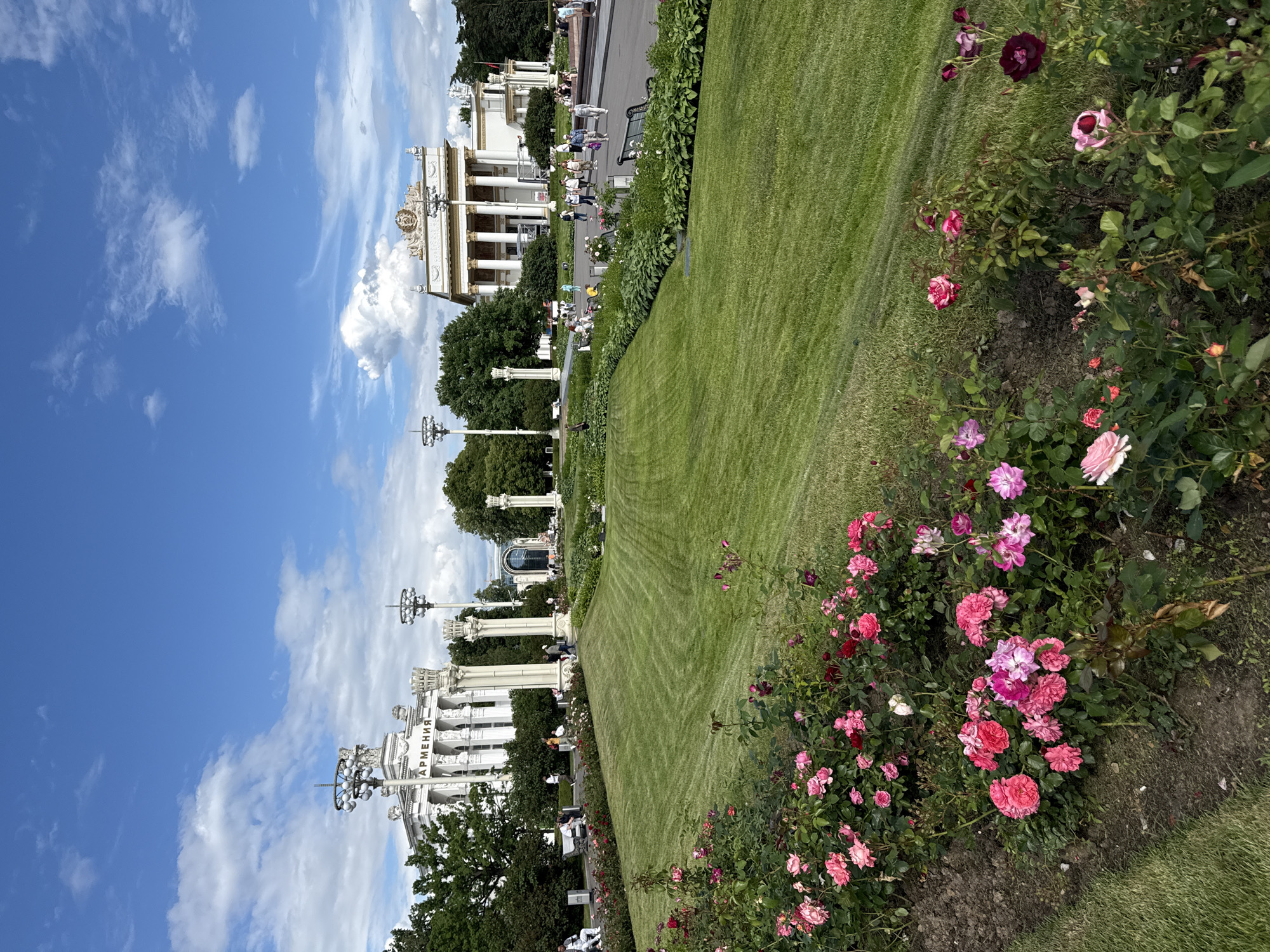 This image depicts a bright, sunny day at an outdoor exhibition or park in Moscow, Russia. The foreground features a vibrant rose garden in full bloom, with a variety of pink and deep red roses adorning lush green bushes. Beyond the flowerbeds, a meticulously manicured lawn stretches into the midground, bordered by a pathway where several people are casually strolling and sitting on benches.
In the background, grand white buildings with classical architectural elements, such as columns and ornate decorations, are visible. These structures appear to be exhibition pavilions, with one clearly displaying the word "АРМЕНИЯ" (Armenia) in Cyrillic lettering. Tall, decorative lampposts stand in front of the buildings. The sky is a clear blue with scattered white clouds, suggesting fair weather and likely mid-day. The overall atmosphere is one of leisure and enjoyment, with people relaxing and taking in the sights of the expansive grounds and impressive architecture.