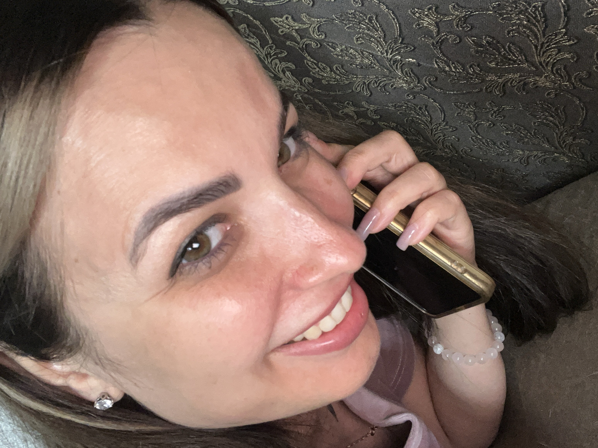 A woman is holding a gold-colored smartphone to her ear and speaking. She has brown hair, brown eyes, and is wearing a light pink t-shirt and a beaded bracelet. The background appears to be a patterned couch.
