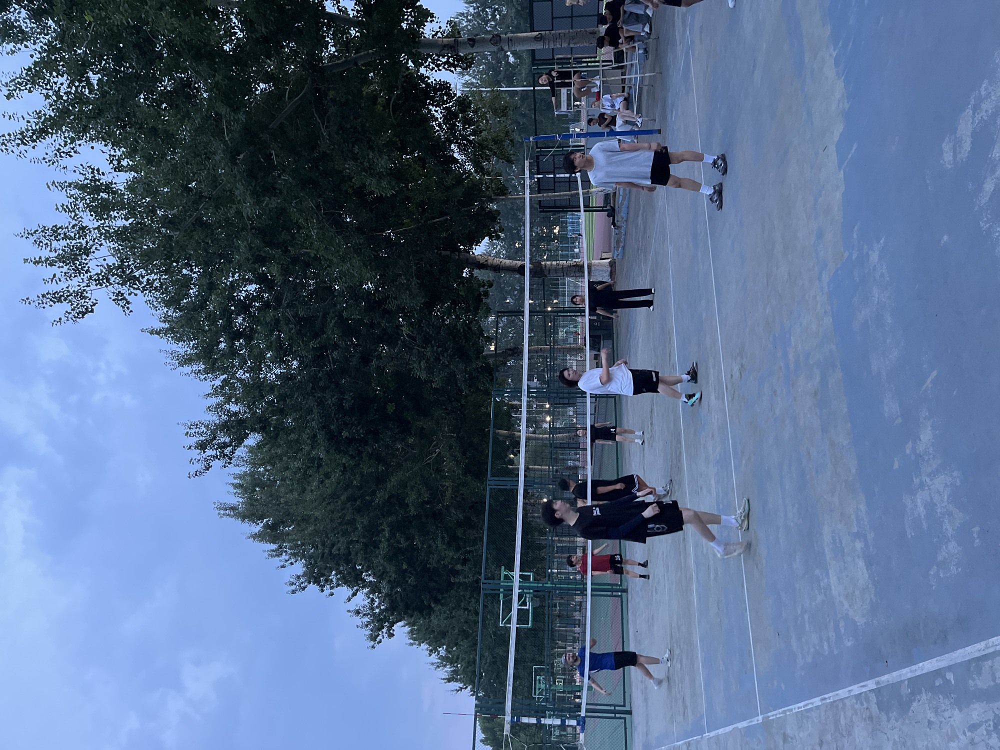A group of young men are playing volleyball on an outdoor court at dusk. The court is blue with white lines. A net divides the court. There is a large tree with dark green leaves on the left side of the image. The sky is a pale blue with some clouds. Some spectators are visible in the background. The people appear to be dressed in casual athletic wear. The overall mood is relaxed and energetic. The time of day is late afternoon or early evening, as indicated by the soft lighting. The location is likely a park or recreational facility in Daxing District, China, given the context.