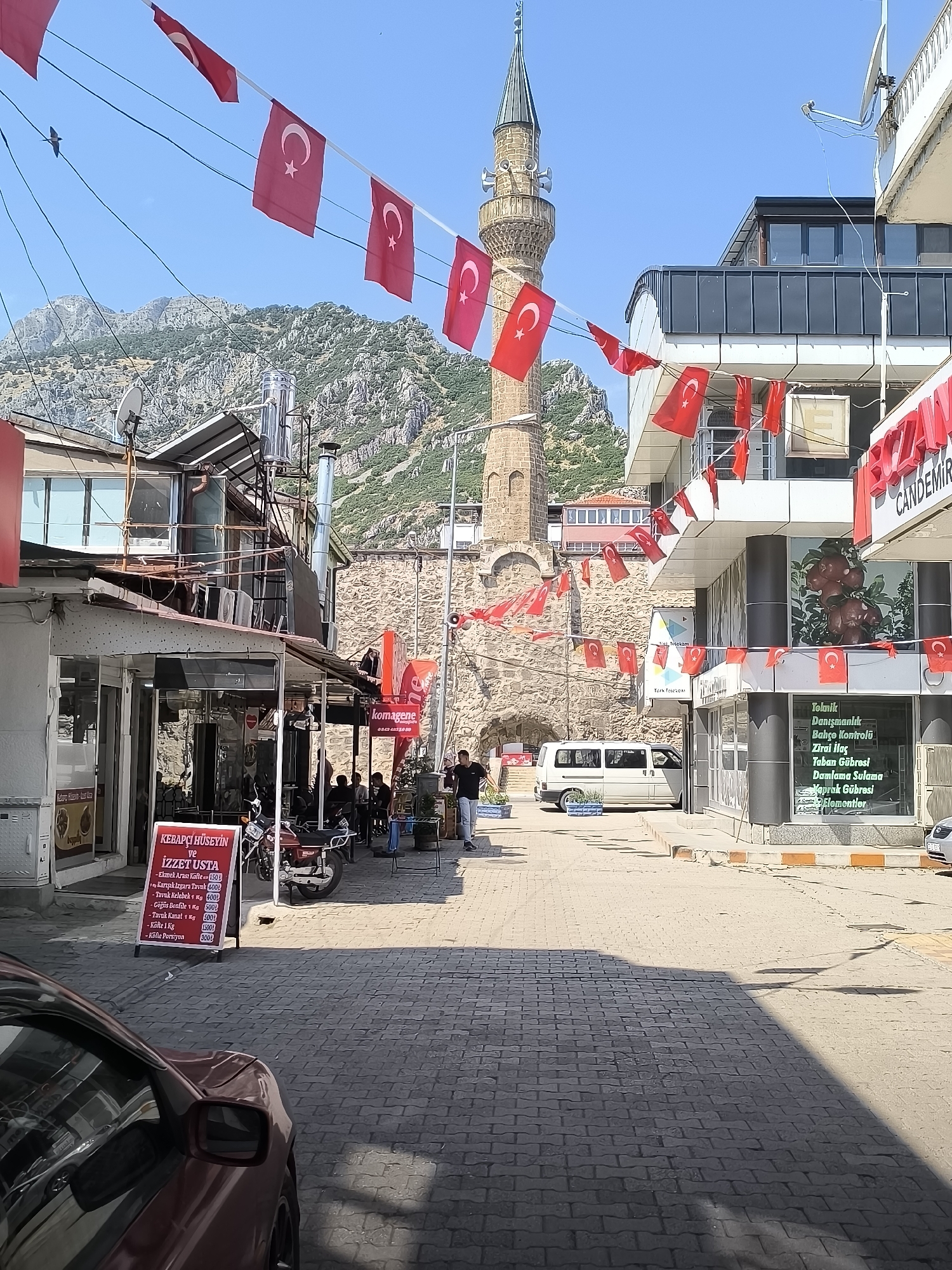 The image depicts a street scene in Eğirdir, Turkey, adorned with numerous Turkish flags strung across power lines. A prominent stone minaret, part of a mosque, rises against a clear blue sky, with rugged mountains in the background. The street is paved with cobblestones and lined with buildings. On the left, a restaurant with outdoor seating displays a menu. Further down the street, a white van is parked. On the right, several commercial establishments are visible, one featuring a sign for "CANDEMIR" and another with window text detailing agricultural products. People are seen walking and sitting at tables, engaged in various activities. The scene is captured under bright daylight, casting distinct shadows.