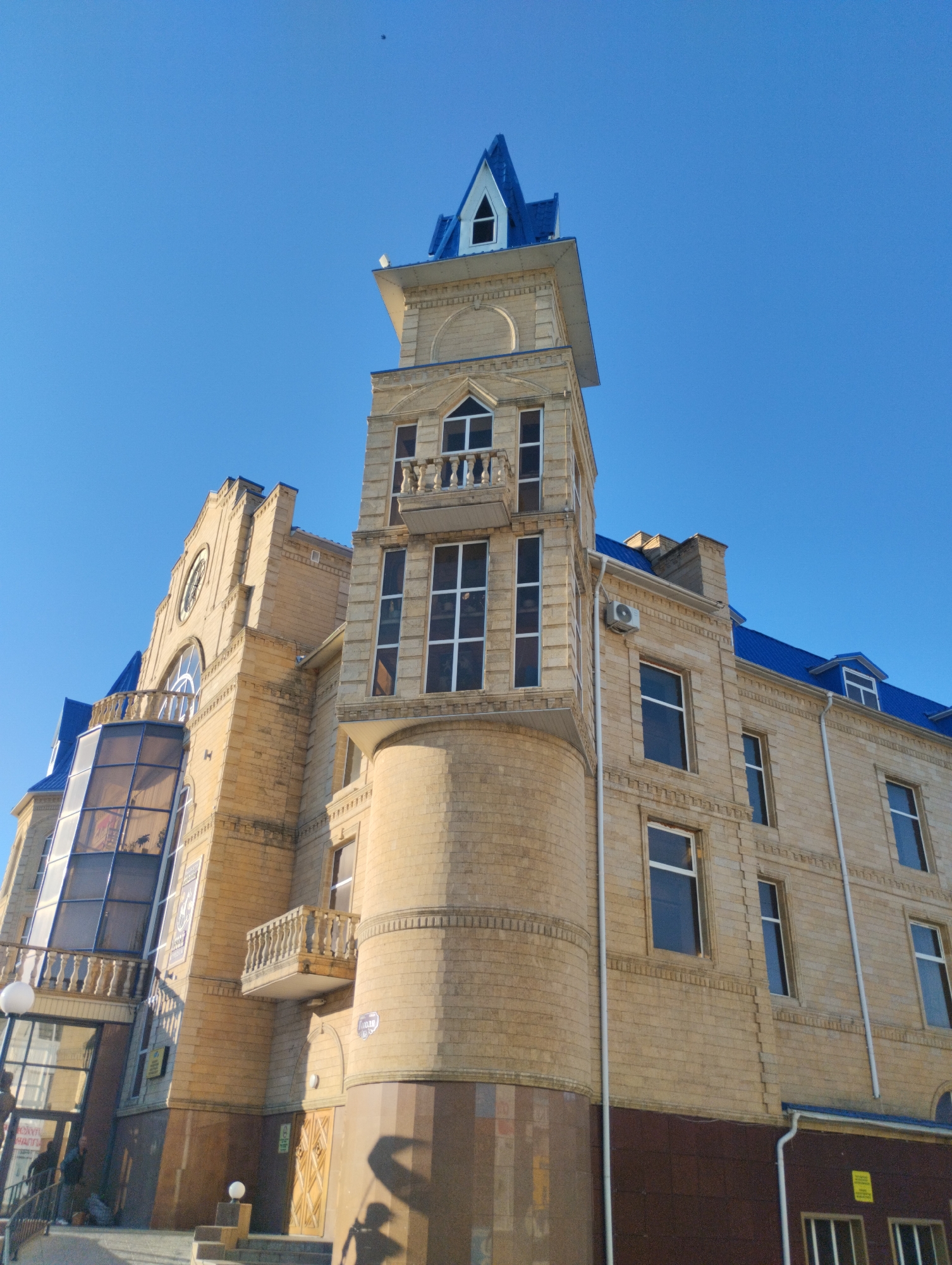 A low-angle, eye-level shot captures a tan brick building with a blue pitched roof against a clear blue sky. The building features architectural details such as a central tower with a pointed blue roof, balconies with balustrades, and numerous windows. Some windows are large and rectangular, while others are arched or have decorative detailing. An air conditioning unit is visible on the side of the building. At the bottom of the image, a glass-enclosed entrance with a railing and a sign with Cyrillic text is partially visible. Shadows of people and a bicycle are cast on the building's facade. The context suggests this is a building in Kostanay, Kazakhstan, given its architectural style and signage.