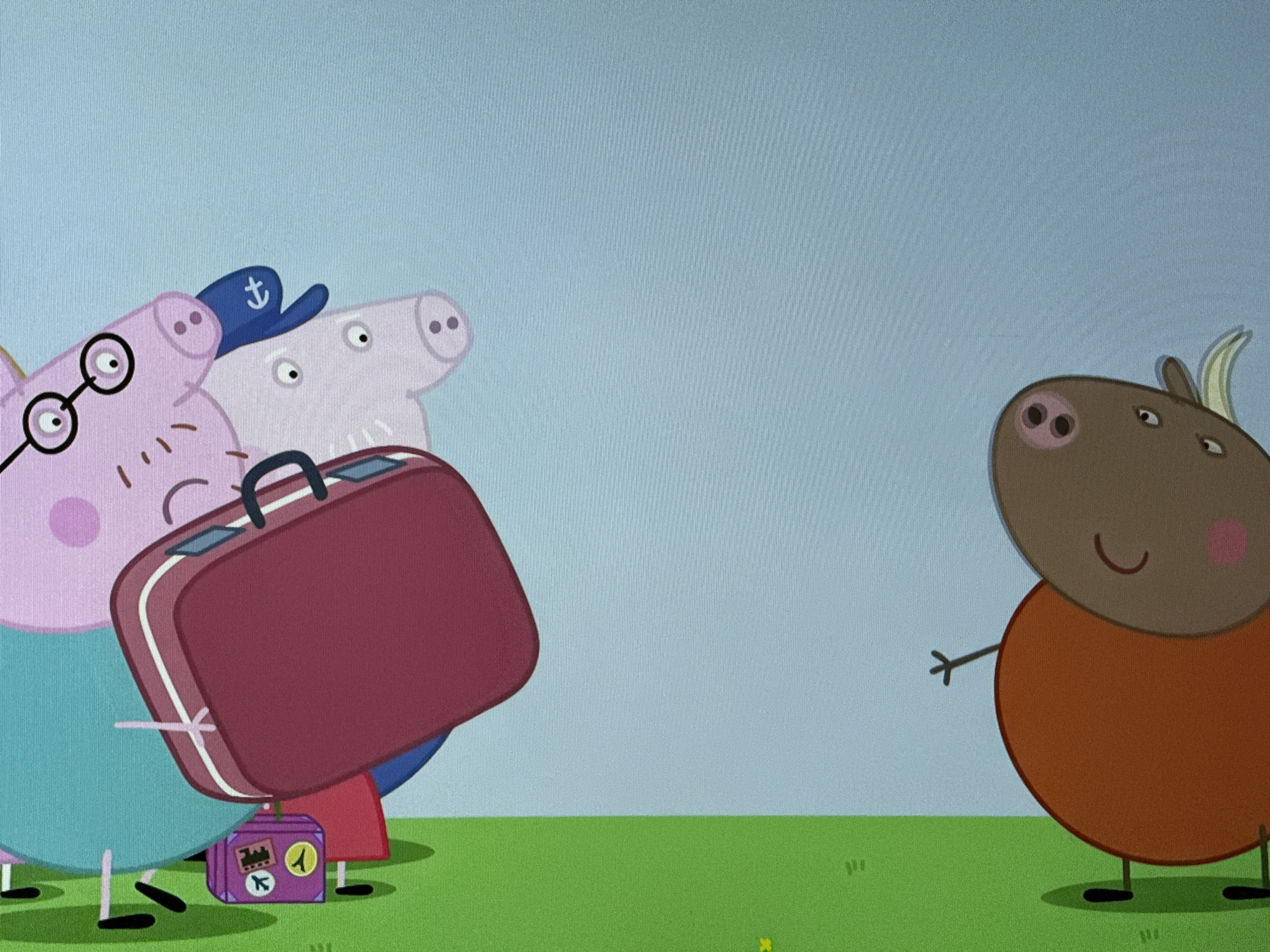 The image is a still from the animated children's show "Peppa Pig." Three pig characters are on the left, one wearing glasses, another a sailor hat. They are carrying a large maroon suitcase and a smaller purple suitcase with travel stickers. On the right, a brown cow character with a simple smiling face is standing with one arm raised in a greeting gesture. The background is a pale blue sky and green grass. There is no indication in the image that it is located in Hazlemere, United Kingdom.