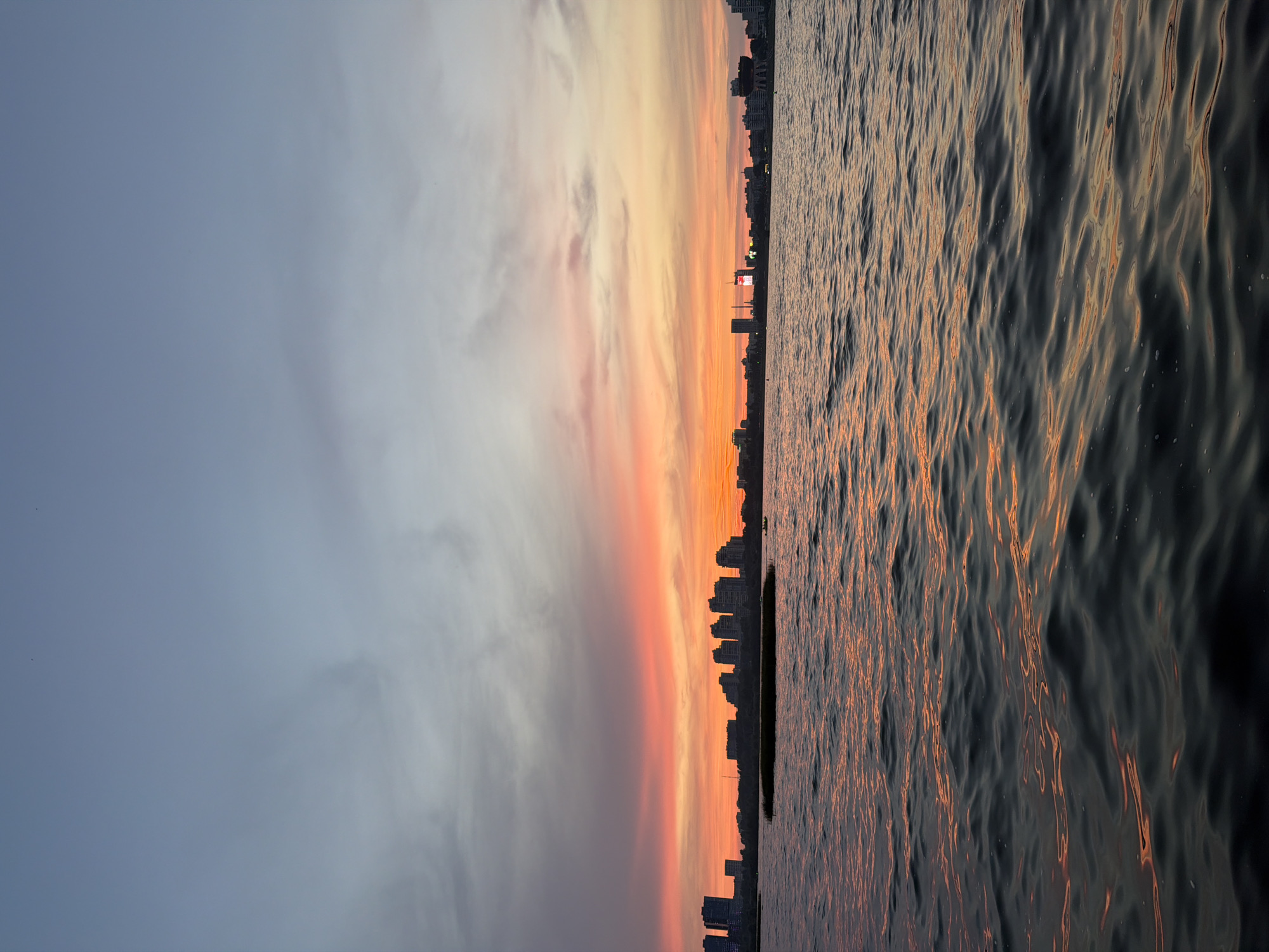 A wide shot captures a body of water reflecting the vibrant hues of a sunset. The sky transitions from a pale blue at the top to a fiery orange and pink closer to the horizon, where a city skyline is silhouetted. The water's surface is rippled, with streaks of orange light from the sunset dancing on the dark waves. Buildings of varying heights form the urban landscape, with some lights visible in the distance. A low, dark strip of vegetation is visible in the mid-ground, separating the water from the city. The overall impression is one of serene natural beauty meeting urban development at the end of the day.