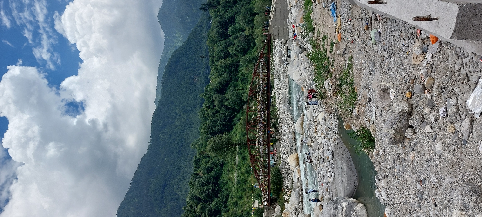 This is a vertical, eye-level, full shot of a river flowing through a rocky terrain with a bridge in the background, under a partly cloudy sky. The scene is set in the city of Kalath, India. The river, with its clear, turquoise water, cascades over smooth, grey and white stones. On the riverbanks, more grey and white stones are scattered, with some patches of green vegetation. A rusty, brown metal bridge spans across the river in the mid-ground, adorned with numerous prayer flags in various colors, flapping gently in the breeze.
On the left side of the riverbank, several individuals are visible. A person in a blue shirt and dark pants stands near the water's edge. A group of people are gathered on the rocks in the river, some sitting, others standing. Further to the right, on the same riverbank, three people are clustered together, one in a pink outfit and two in lighter colored clothing.
The background features a densely forested mountainside, covered in dark green trees that extend up to the crest of the mountain. The sky above is a mix of bright blue patches and large, fluffy white clouds, with some areas of darker, heavier clouds suggesting potential rain. In the foreground, the ground is covered in gravel and stones, with scattered debris, including plastic bags and paper waste. A concrete barrier with exposed rebar is visible along the right edge of the frame.