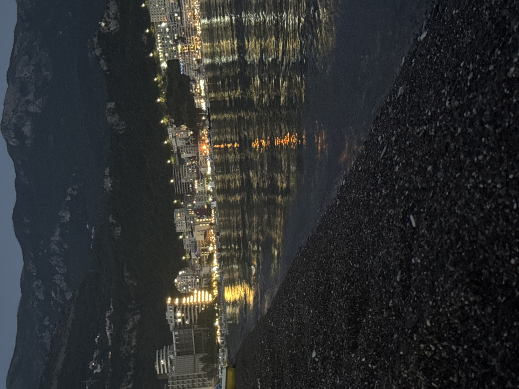 The image depicts a night scene of a coastal city, likely Boreti, Montenegro, viewed from a dark pebble beach. In the foreground, the beach is composed of small, dark pebbles with some lighter-colored stones scattered throughout. The sea, in the middle ground, reflects the lights of the city and the sky, creating shimmering patterns. The water appears calm with gentle ripples.
Beyond the water, the city skyline is illuminated with numerous lights, indicating it is nighttime. Buildings of various heights and styles are visible, with many windows lit up. Some of these buildings appear to be hotels or apartment complexes, with a festive, urban atmosphere. The lights suggest a lively environment.
In the background, dark, forested mountains rise steeply, their contours softened by the dim lighting. The sky above is dark, characteristic of nighttime, with a subtle gradient suggesting the fading twilight. The overall impression is one of a picturesque coastal city at night, with the natural beauty of the mountains and sea complementing the urban landscape. No people are visible in the frame. The image has a slightly grainy texture, common in low-light photography.