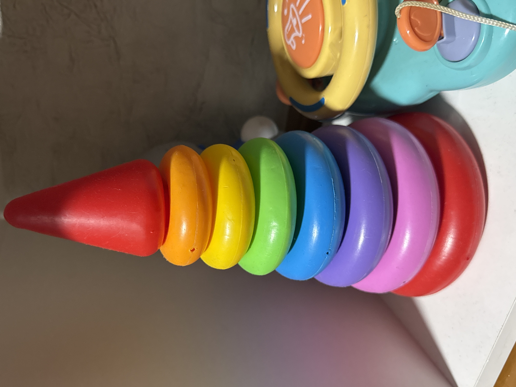 In the city of Yarominski Sel'skiy Sovet, Belarus, a close-up, eye-level, studio shot captures a brightly colored children's toy. Dominating the frame is a stacking ring toy, composed of several concentric rings in various hues of the rainbow: red, orange, yellow, green, blue, purple, and pink. These rings are stacked in descending order of size, culminating in a red cone at the apex. To the right of the stacking toy, a portion of another child's toy is visible; this toy is turquoise with yellow accents and features a circular steering wheel-like component and an orange button with a bell icon. The background is softly blurred, featuring a textured, neutral-toned wall and a white surface in the foreground, suggesting an indoor, possibly domestic or retail, setting. The lighting is even, highlighting the smooth, plastic texture of the toys. There are no people present in the image, and no actions are taking place.
