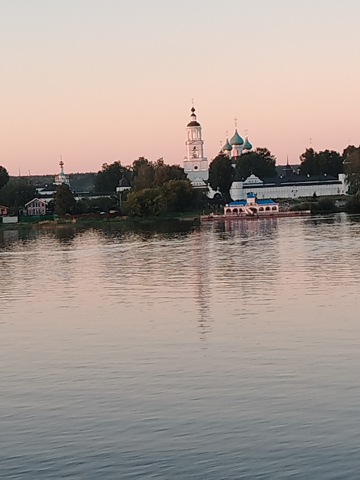 The image presents a wide-angle view across a body of water towards an architectural complex situated on the far bank in the city of Заволжье, Russia. The sky above is clear, displaying a gradient of pale pink transitioning to a lighter shade upwards, indicative of either sunrise or sunset.
The foreground is comprised of the calm surface of a wide river or lake, exhibiting subtle horizontal ripples. The water's surface reflects the light tones of the sky and distant landscape.
On the distant bank, a line of green foliage, including trees and bushes, separates the water from the structures. The central feature is a large, white-walled complex. A tall, multi-tiered white bell tower with arched apertures is visible on the left-center, capped by a dark, pointed spire and a golden onion dome adorned with a cross. To the right and slightly behind the bell tower, several white buildings with distinct teal-colored onion domes, each topped with a golden cross, mark the primary religious edifices within the complex. These structures are encompassed by a robust white wall.
Further left on the distant bank, a separate multi-story white building with a dark roof is partially obscured by trees. Directly in front of the main complex, extending into the water, is a white structure with a blue roof and multiple arches, which could be a chapel, gazebo, or pier, connected to the shore.
No human figures, discernible actions, or interactions are present in the scene. The overall environment conveys a peaceful, unpopulated setting with historical architecture.