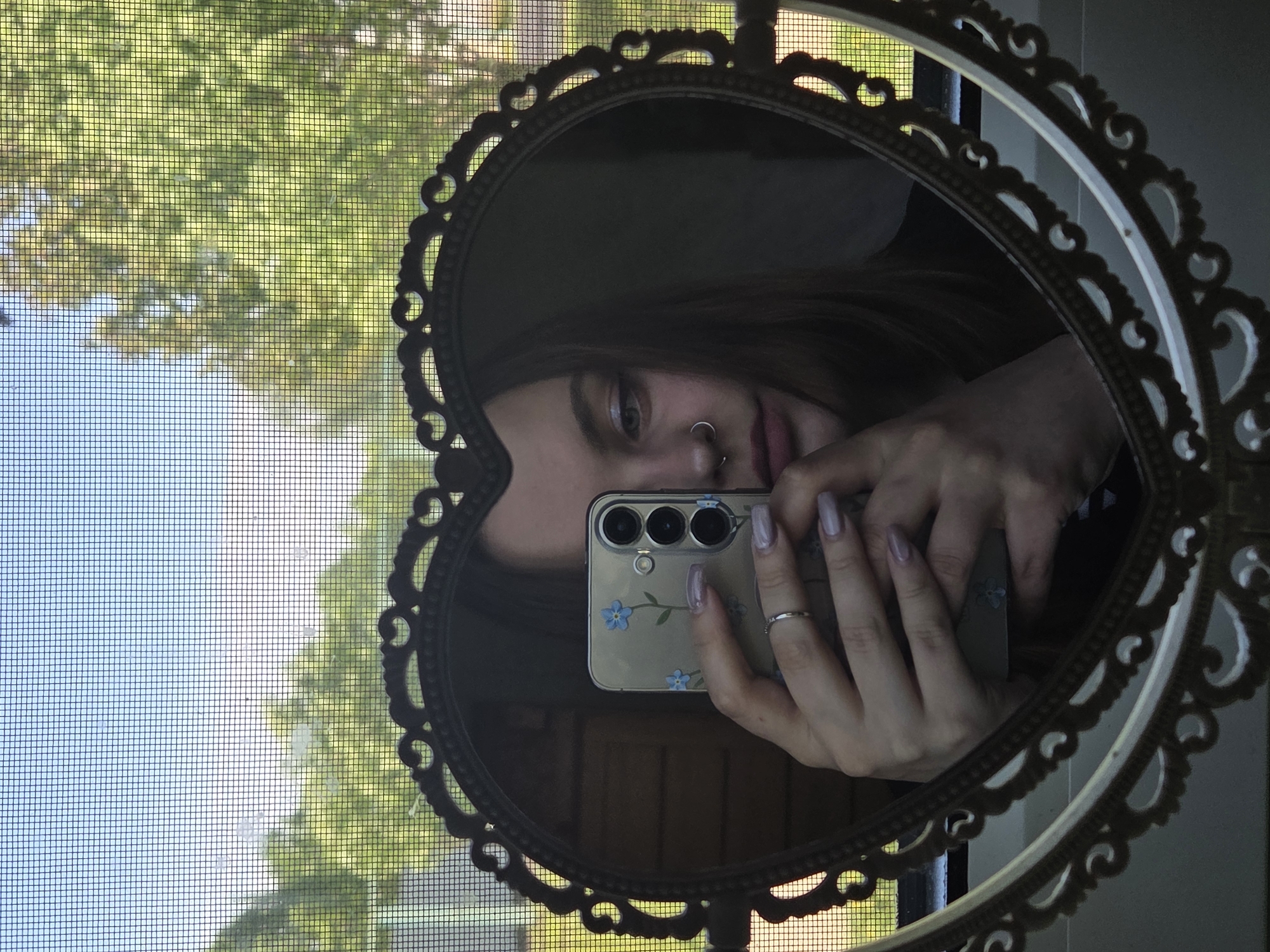 The image shows a selfie taken in a heart-shaped mirror. A person is holding a smartphone up to the mirror, capturing their reflection. The phone has a transparent case with blue flower decorations. The person has pierced nostrils and wears a ring. The reflection includes part of the person's face and hand. Outside the window, visible through the mesh, are trees. The location and specific city are not discernible from the image.
