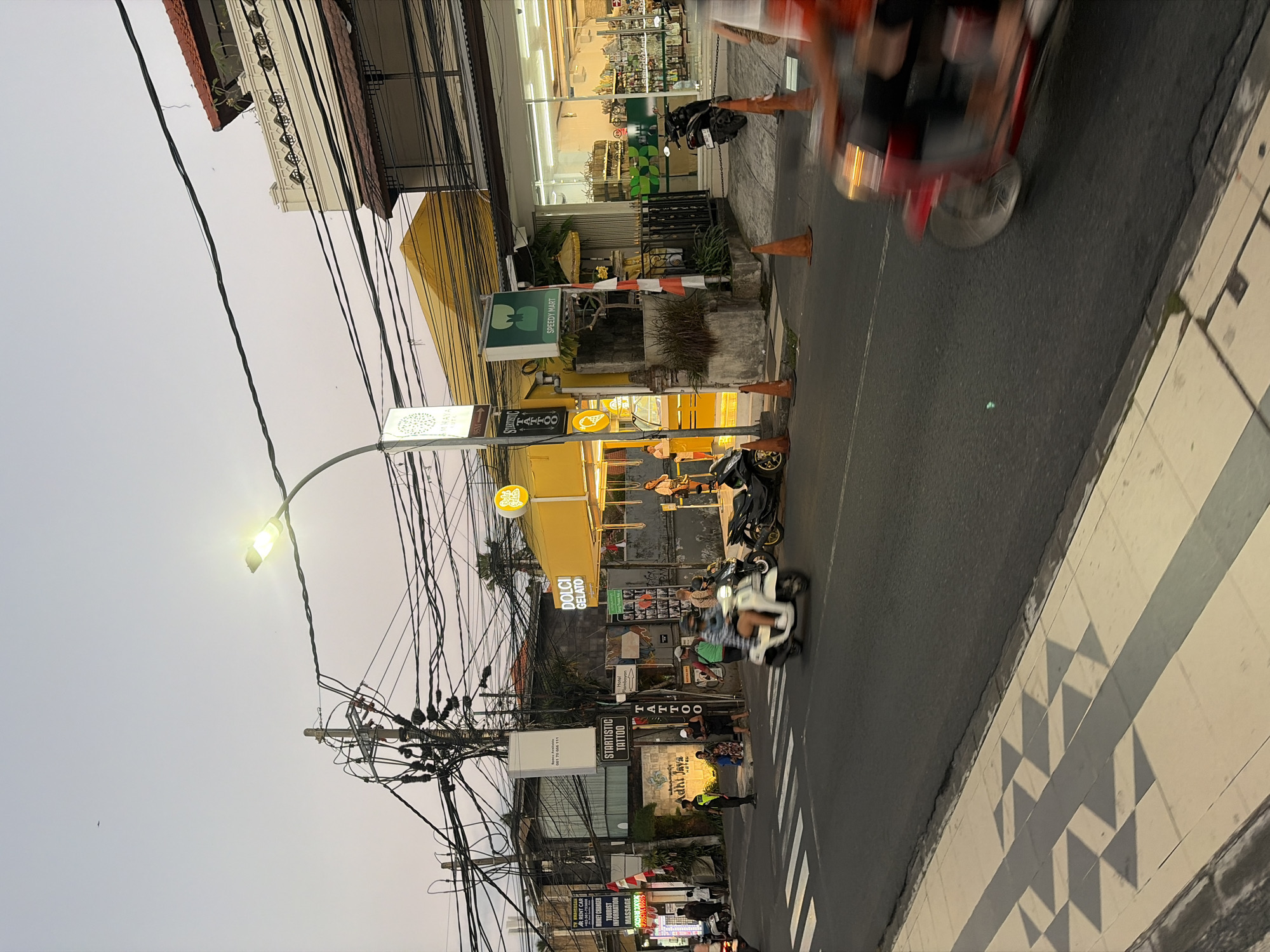 This is a street scene in Kuta, Indonesia, during dusk. The street is paved with asphalt and has a pedestrian walkway with a patterned tiled surface. Several buildings line the street, including a yellow building with "DOLCI GELATO" signage, a building with a white facade and ornate detailing, and a "SPEEDY MART" convenience store with a glass front displaying goods. A prominent feature of the scene is a dense network of electrical wires overhead, supported by utility poles.
On the street, a white scooter is in motion, with a rider visible. Another motorcycle is parked near the Speedy Mart. A blurry, red three-wheeled vehicle, possibly a tuk-tuk, is also in motion in the foreground, its form indistinct due to speed. Several people are visible walking on the sidewalk and near the buildings, some interacting or standing. Orange traffic cones are placed along the edge of the road. The sky is a pale, dusky hue, suggesting the end of daylight. Signs visible include "AMAYA ROTA," "STARTISTIC TATTOO," and a "Hotel Namboyan" sign. The overall atmosphere is that of a busy commercial street in a tropical urban environment.