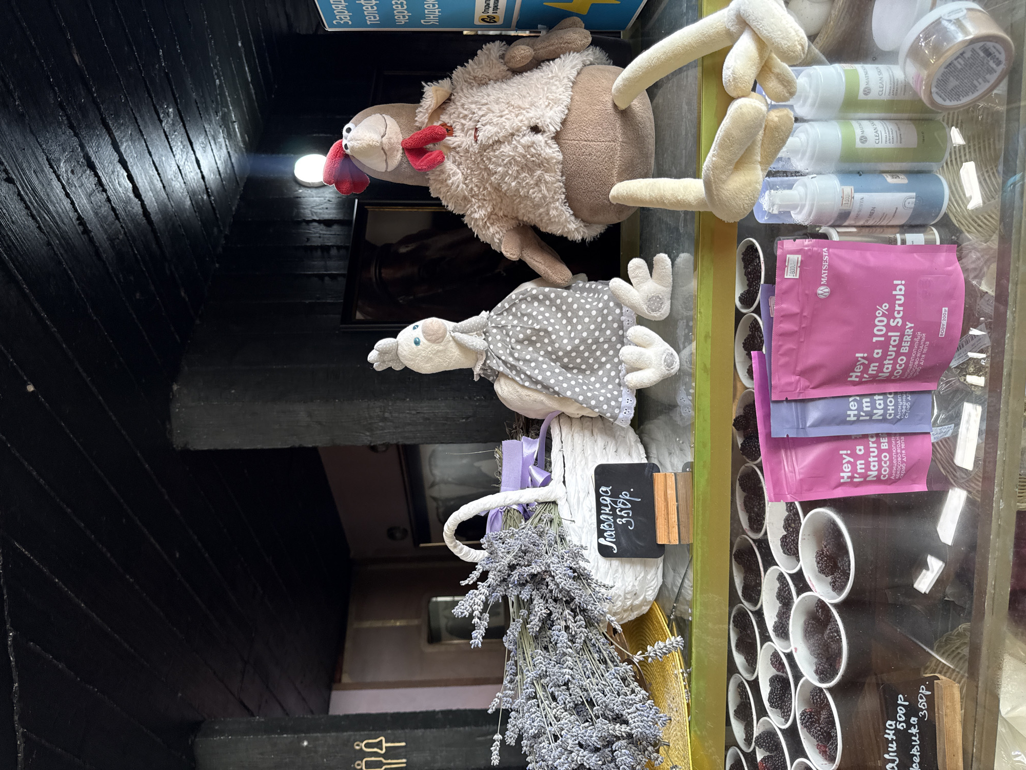The image depicts a store display in Progress, Russia, featuring an assortment of items, including lavender, plush toys, and skincare products. A white basket filled with dried lavender is prominently displayed, accompanied by a small chalkboard sign indicating "Лаванда 350р." (Lavender 350 rubles). Two plush chicken figures, one wearing a gray polka-dot dress and the other a fluffy brown coat, are positioned behind the lavender. In the foreground, several small white cups are filled with blackberries. A selection of skincare products, including pink and purple pouches labeled "Hey! I'm a 100% Natural Scrub!", and several bottles of foaming cleanser, are also visible on the shelf. The background shows a dark wooden ceiling and walls, with a blue sign in the upper right corner featuring Russian text that appears to be related to charging phones or a service offered by Yandex.