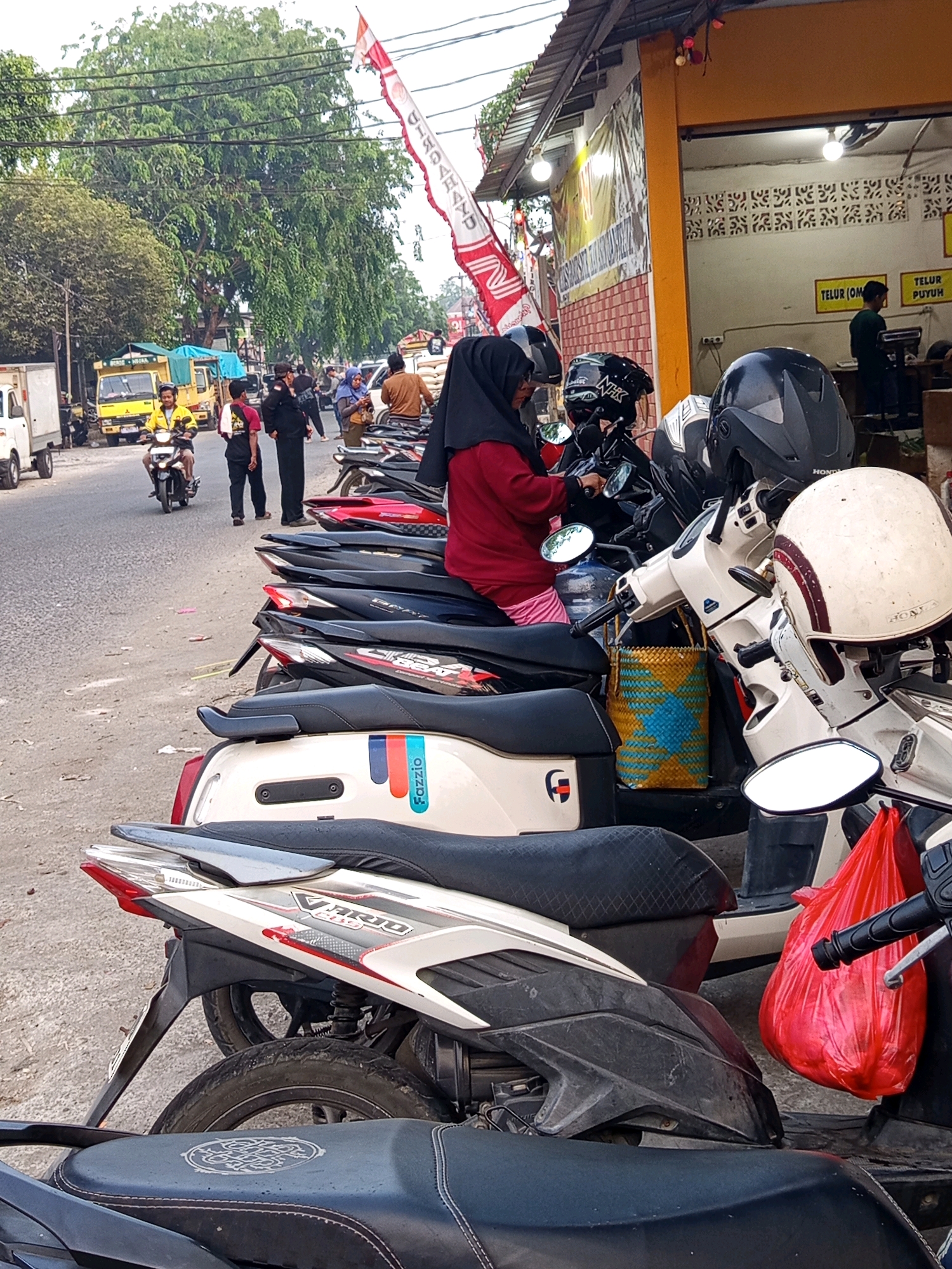 In Karawang, Indonesia, a street scene unfolds with a row of parked motorcycles in the foreground, predominantly scooters, casting shadows on the asphalt. A woman wearing a red hijab and a maroon jacket is seated on one of the scooters, facing away from the camera, while another woman in a blue hijab stands nearby. A parked white truck and a yellow truck with a blue tarp are visible on the street, along with several people walking and standing around, some wearing uniforms. A building with an orange facade and a yellow awning, possibly a shop, is present in the background, with signs in Indonesian indicating "TELUR PUYUH" (quail eggs). A red plastic bag hangs from one of the motorcycles. The overall environment suggests a busy commercial area with ongoing daily activities.
